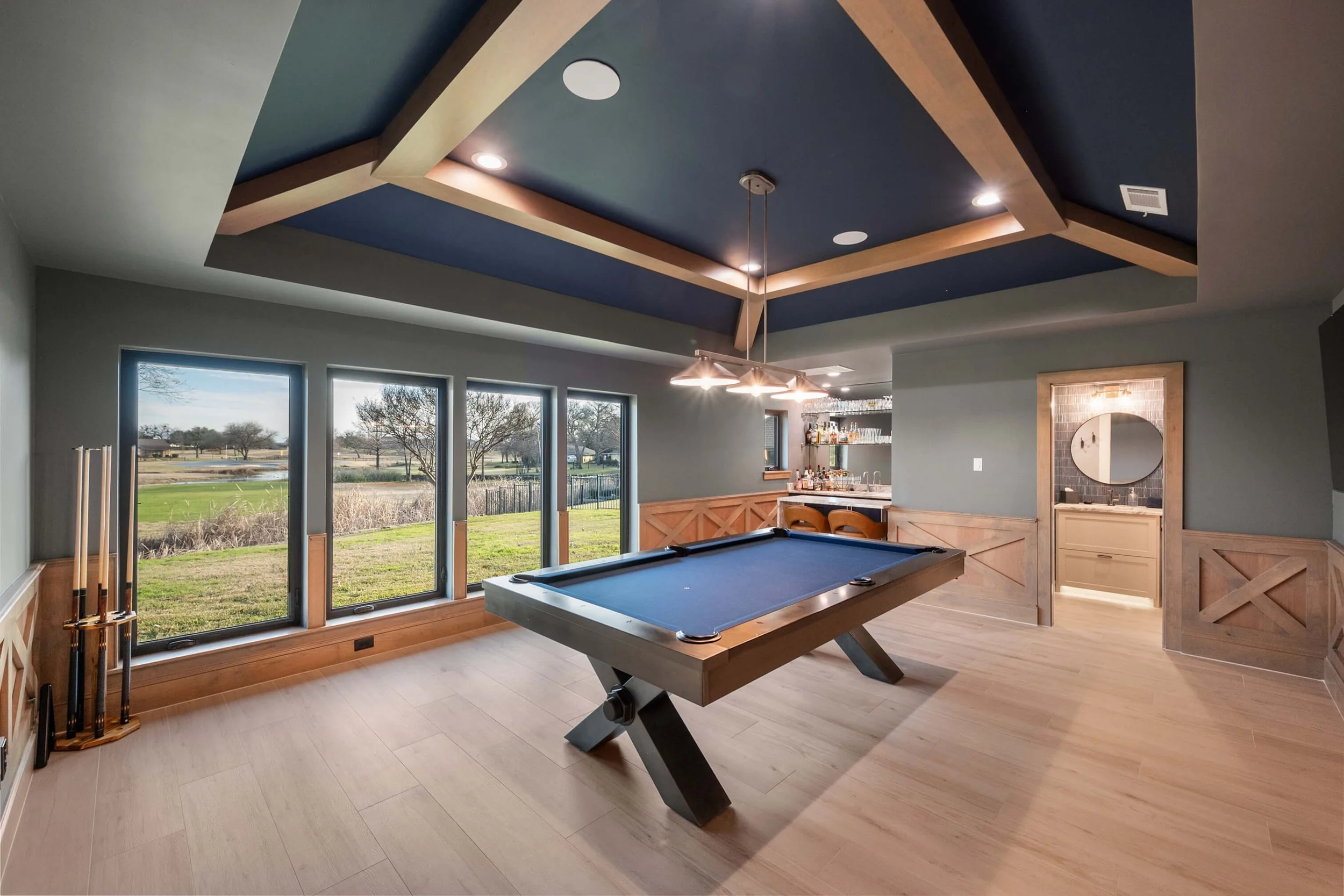 A spacious game room with a pool table, large windows showing an outdoor landscape, a bar area with stools, and a small bathroom with a round mirror. The room features a wooden ceiling with blue accents and recessed lighting.