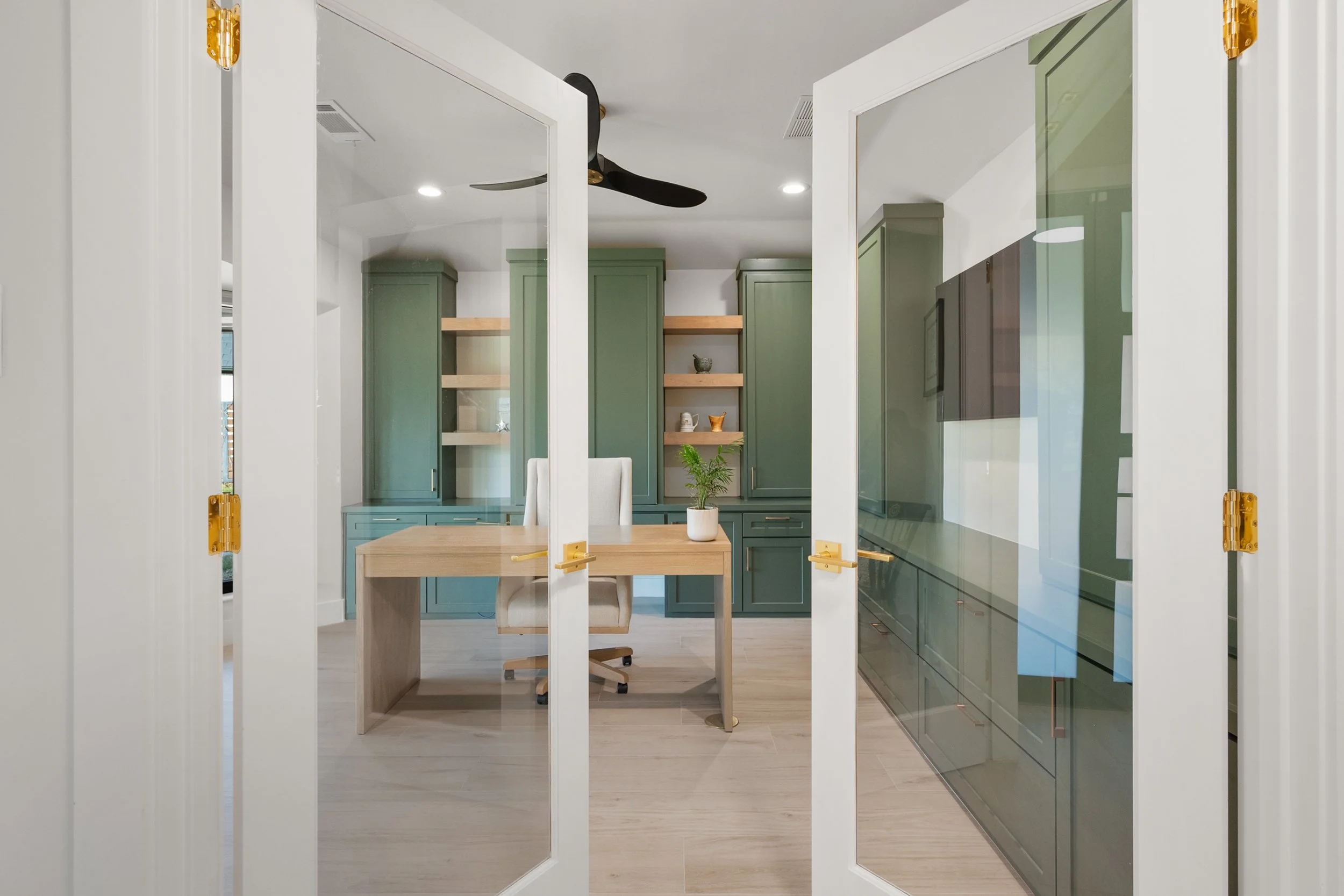 View through glass doors into a home office with green cabinets, a wooden desk, a white chair, and open wooden shelves with decor.