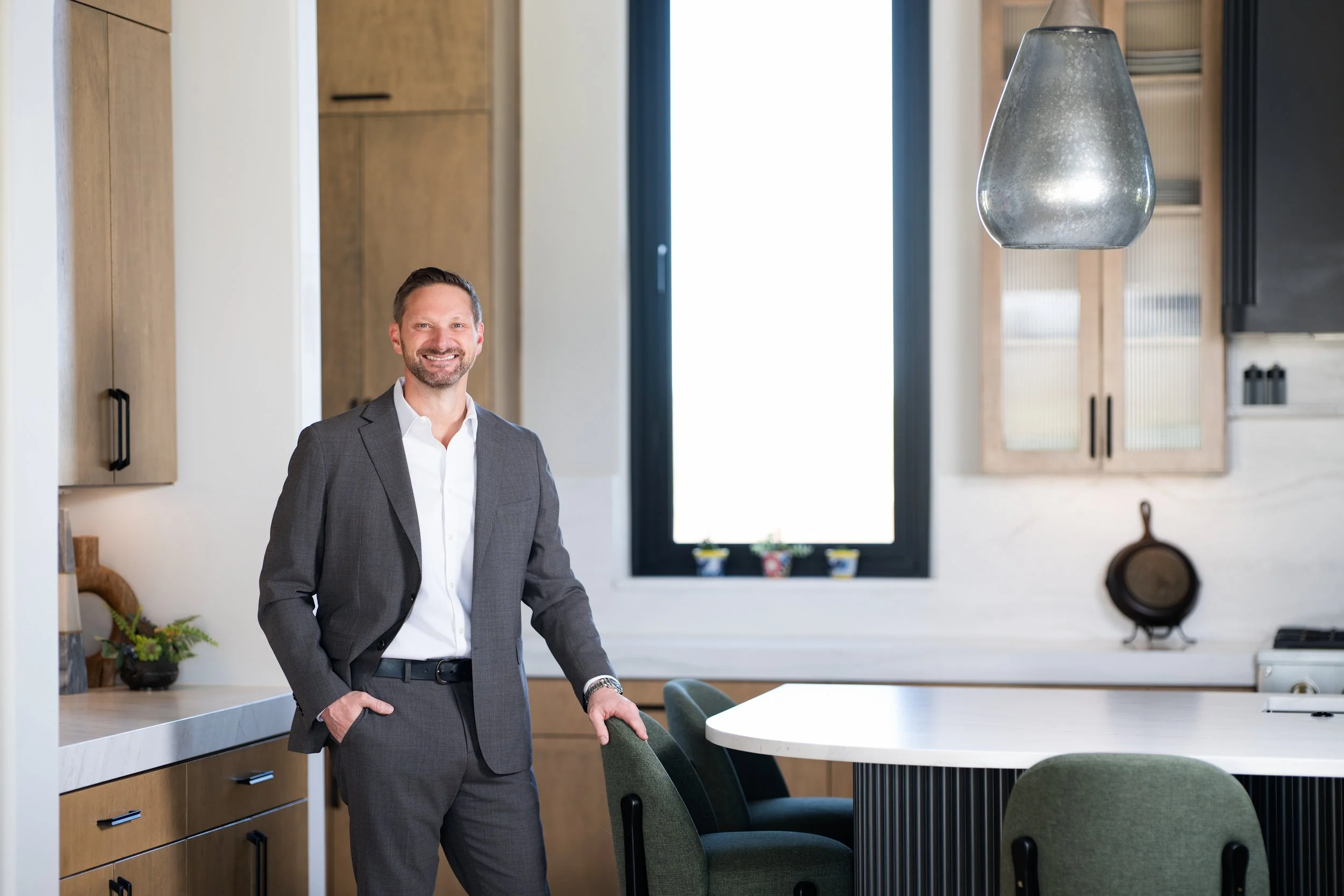 A man in a gray suit smiling, standing in a modern kitchen with wooden cabinets, a white countertop, and a large window with a black frame.