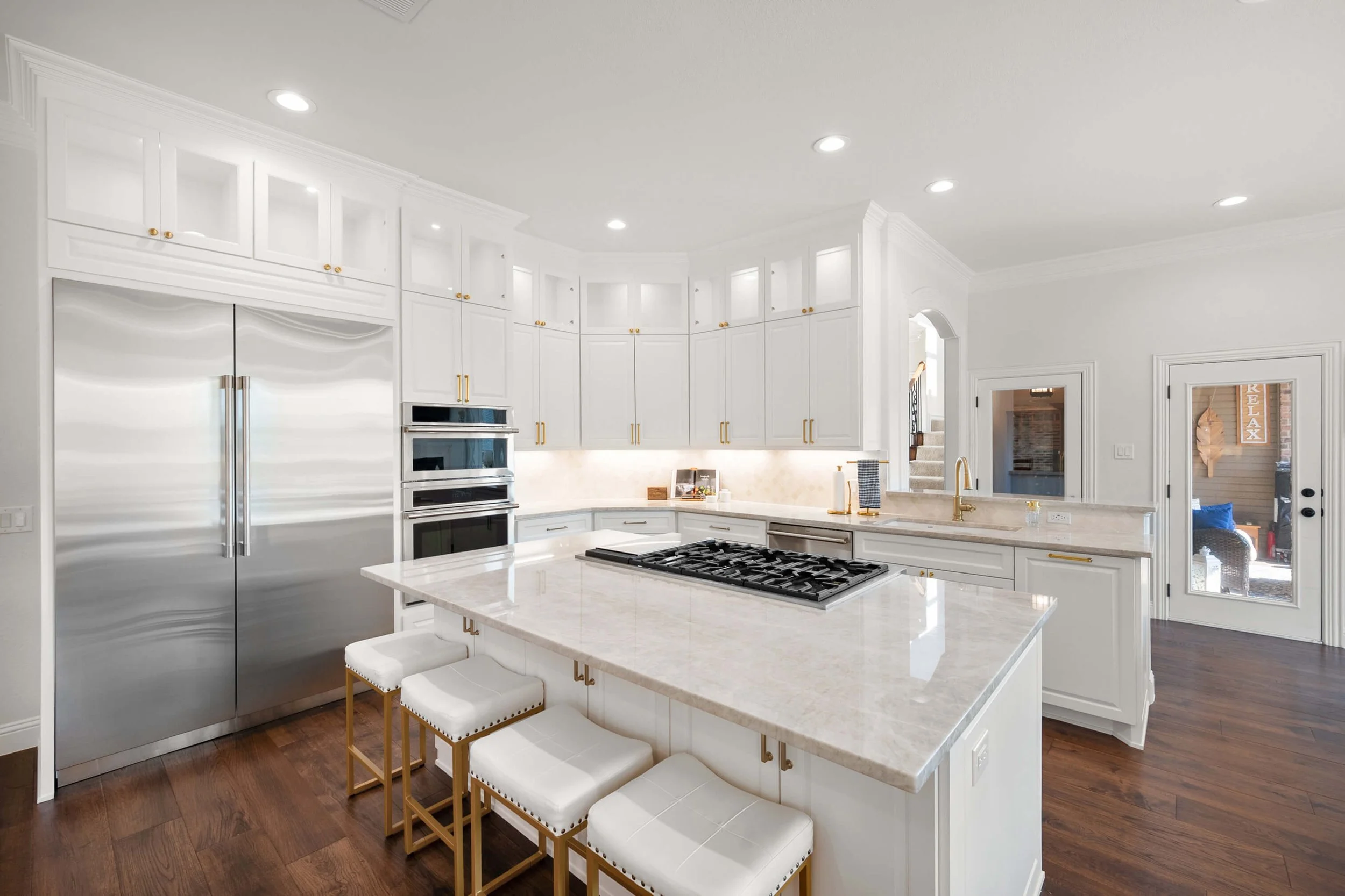 Modern white kitchen with gold hardware, stainless steel refrigerator, and gas cooktop island. Bright lighting and hardwood floors.