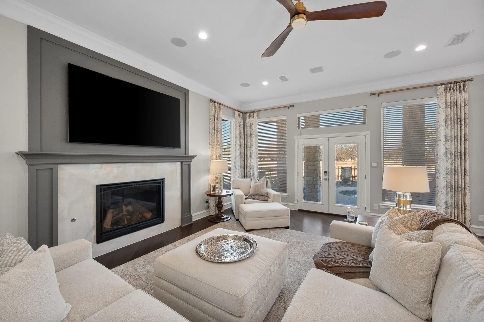 Living room with beige sectional sofa, armchair, wood coffee table, fireplace, large wall-mounted TV, windows with blinds and curtains, ceiling fan, lamps, and area rug.