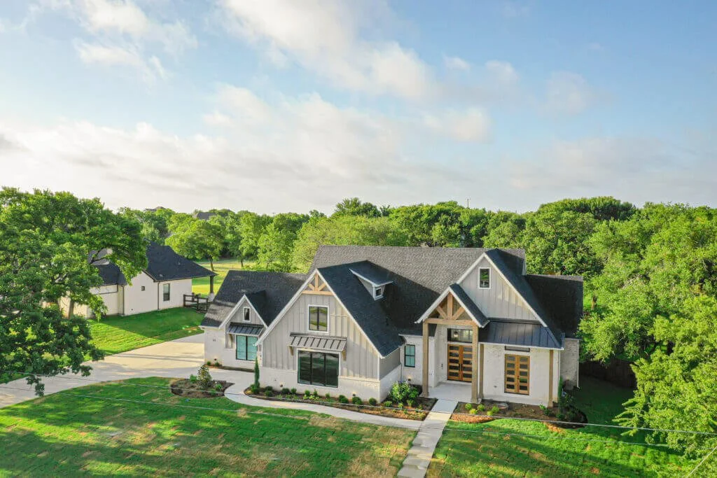 An aerial view of a modern house with a black roof, surrounded by green trees and grass, under a partly cloudy sky in Mansfield, TX.