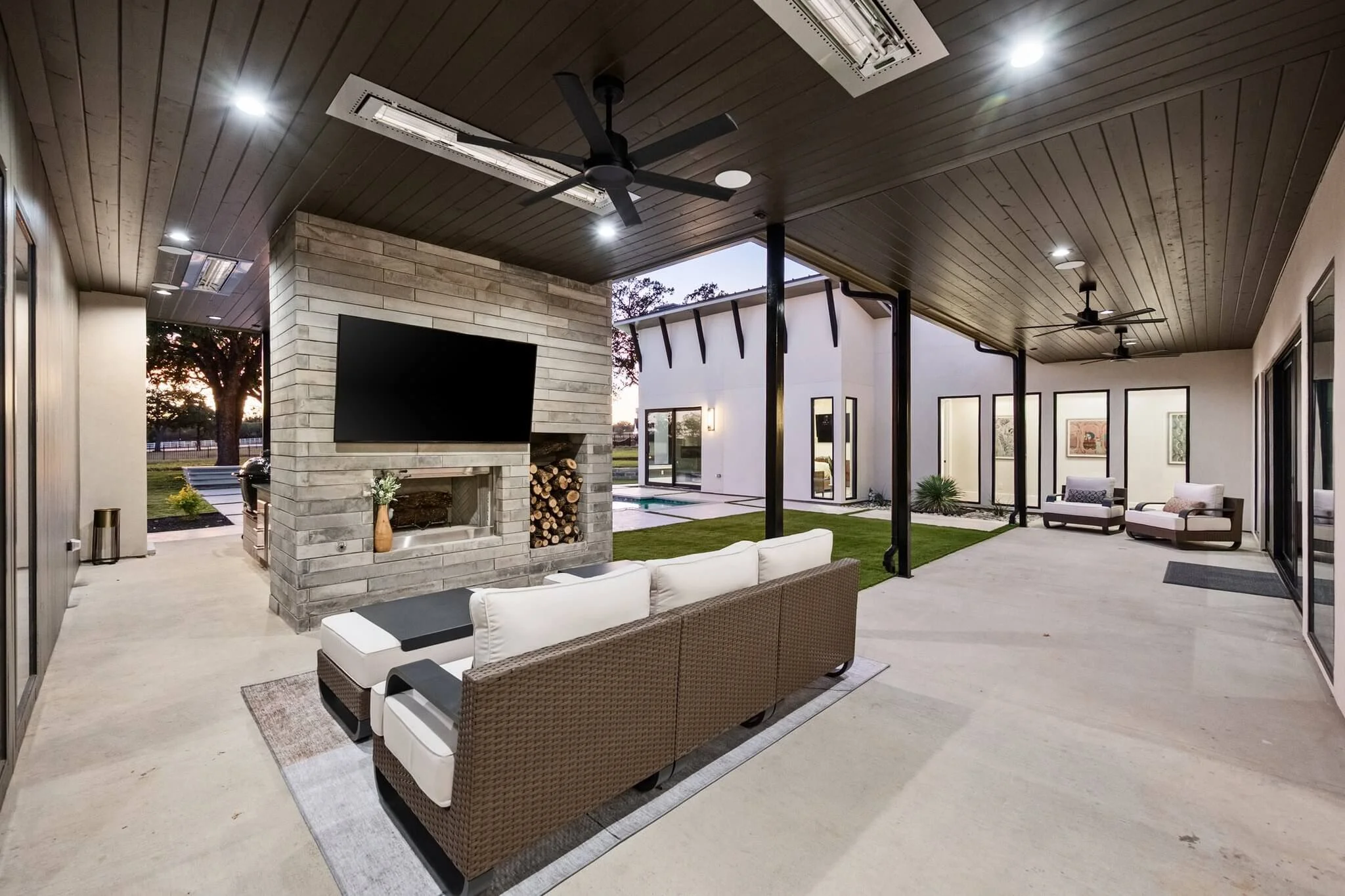 Covered outdoor patio area with ceiling fans, a brick fireplace with wood logs, a large flat-screen TV mounted above the fireplace, and outdoor seating including a sectional sofa and two armchairs. There are skylights in the ceiling, and the space op
