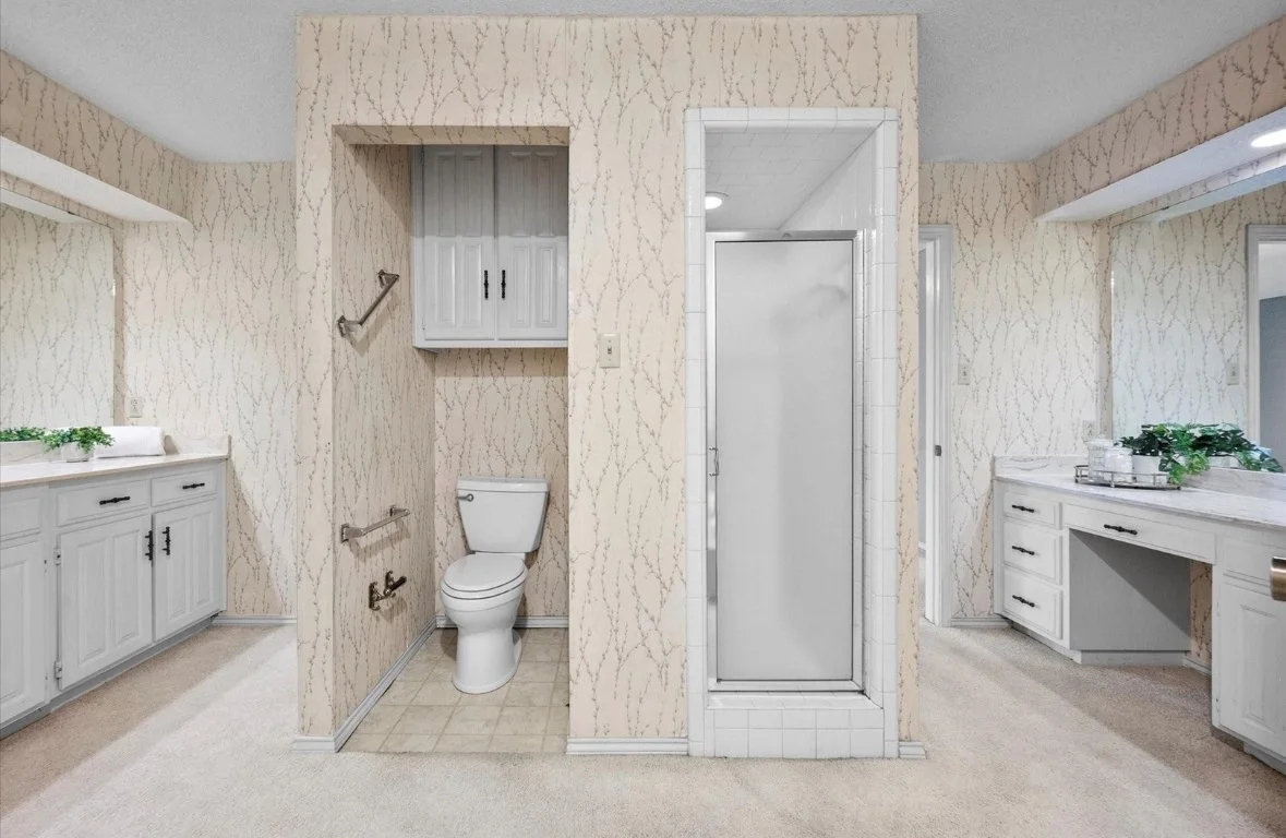 A bathroom with a toilet, two built-in vanities with mirrors, and a shower stall. The walls have beige patterned wallpaper, and the room has white cabinetry and beige carpeting.
