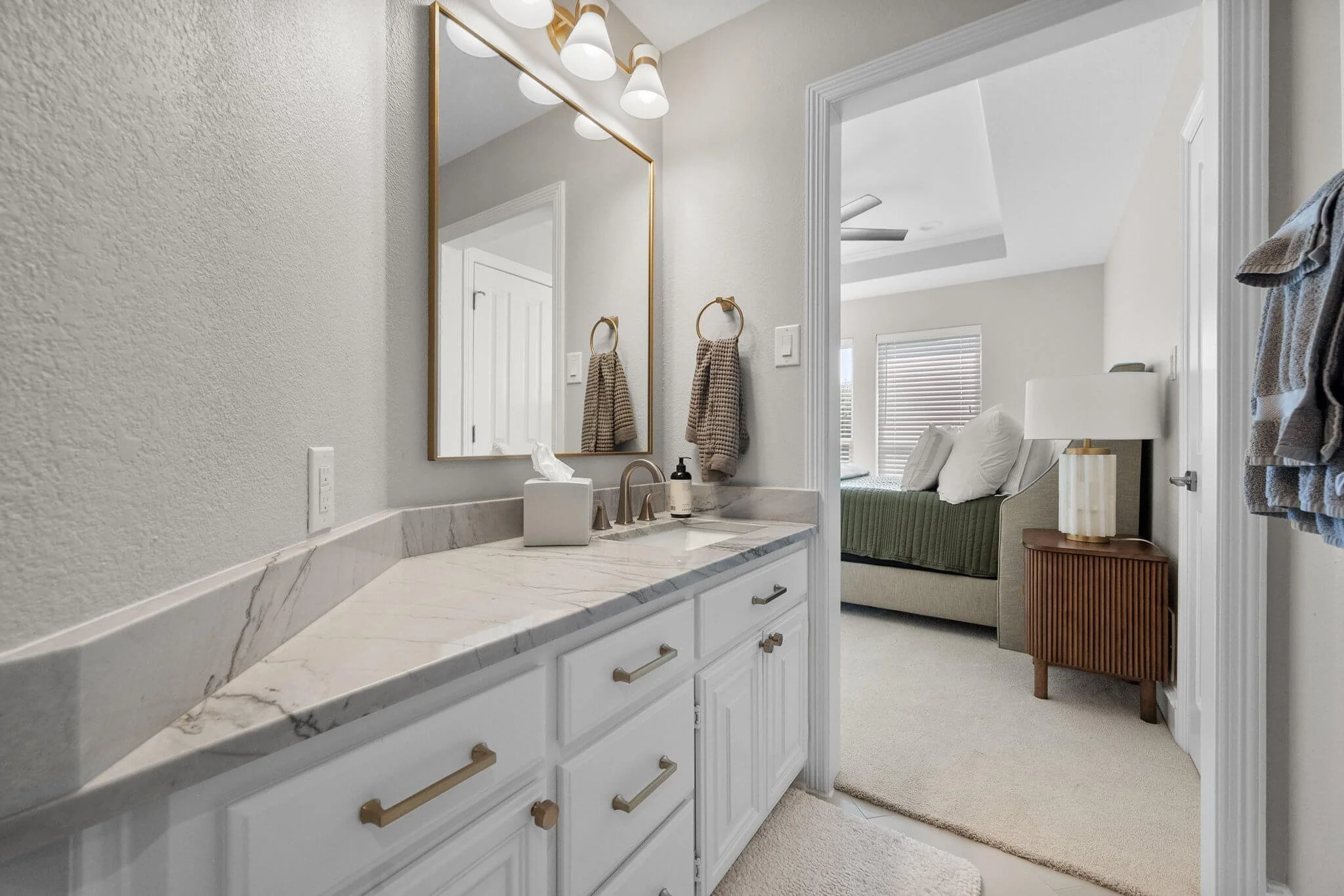 Bathroom vanity with marble countertop, mirror, wall-mounted light fixtures, towels on hooks, and bedroom visible through doorway with bed and nightstand.