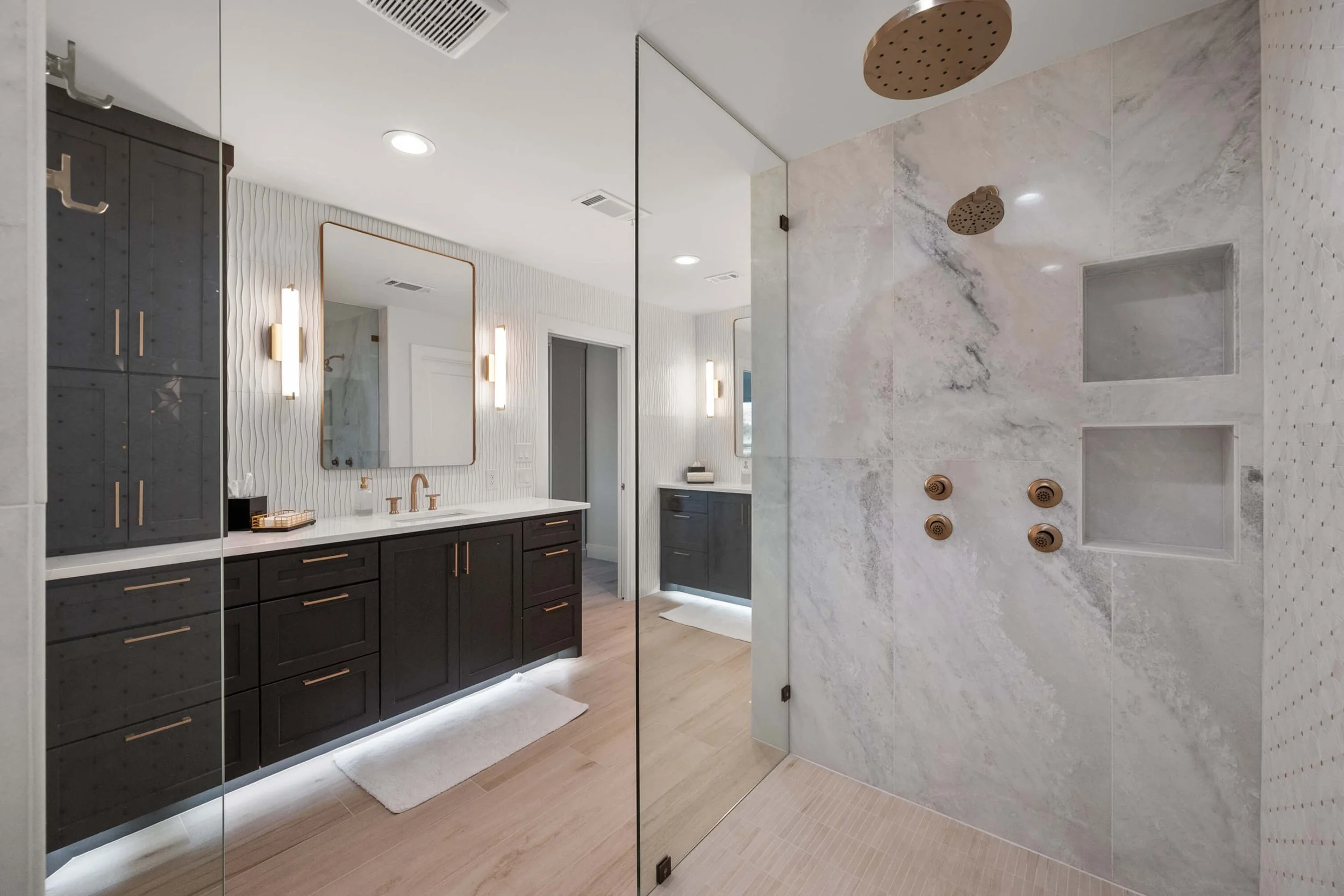 Modern bathroom with a large glass shower enclosure, dark vanity with gold handles, white countertop, mirror, and wall-mounted lights, featuring stone and tile surfaces.