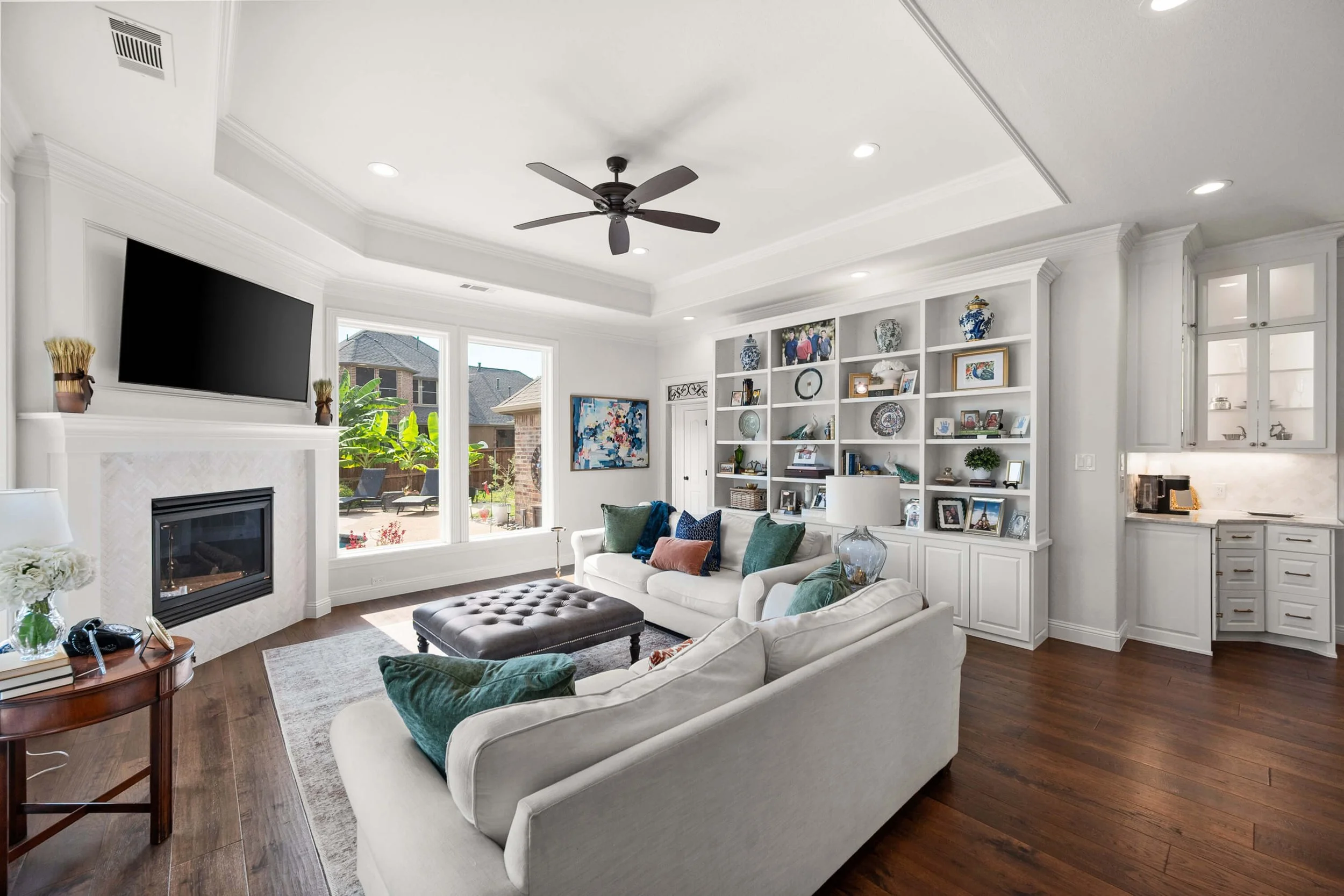 Bright living room with white sofas, a built-in white bookshelf filled with decorative items and photos, a mountain of pillows, a dark brown leather ottoman, a large window with a patio view, hardwood floors, a ceiling fan, recessed lighting, a wall-