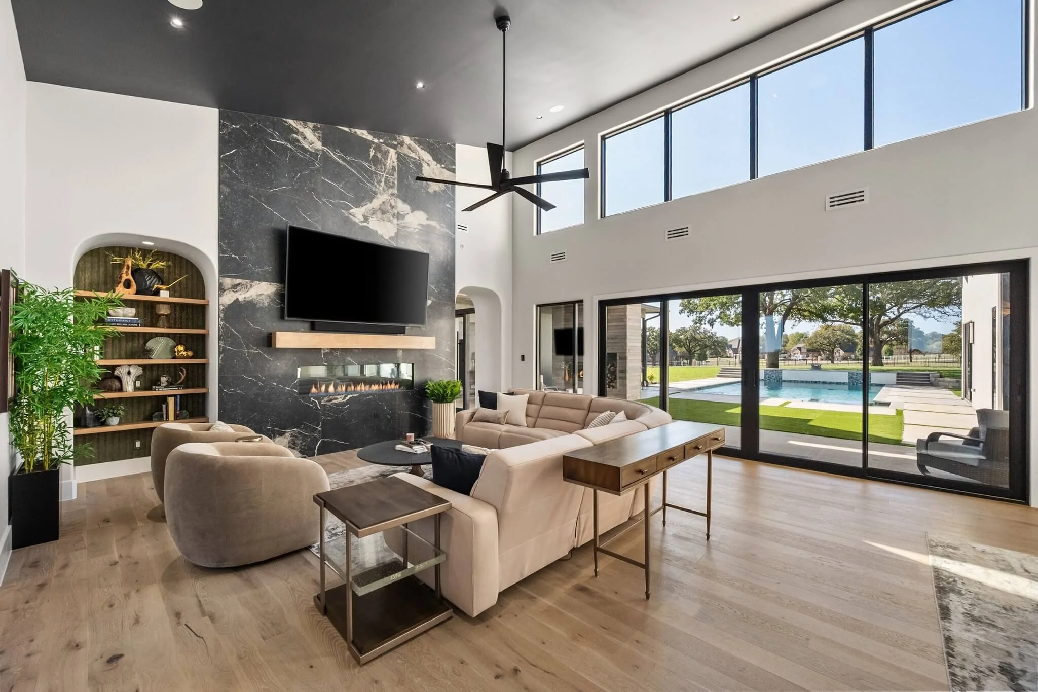 Modern living room with large glass sliding doors leading outside to a pool and yard, featuring beige seating, a black marble fireplace, a large flat-screen TV, and wooden flooring.