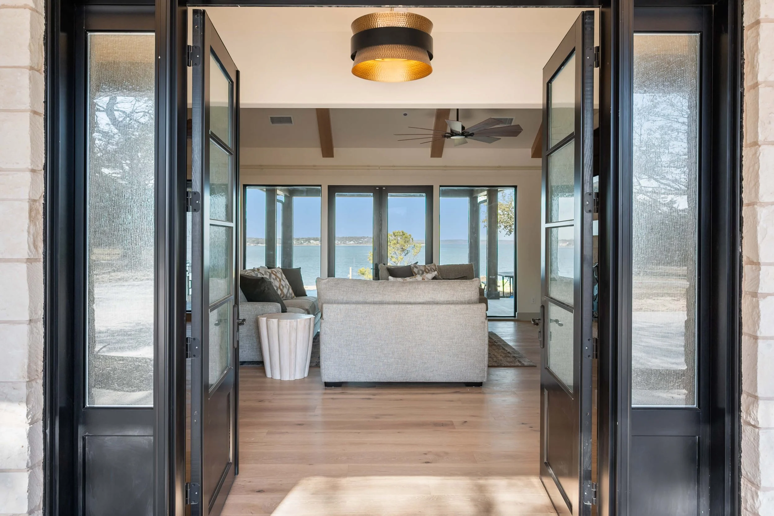 Open black-framed glass doors leading into a living room with view of water through large windows, featuring sofas, a side table, and ceiling fan built by Lakeland Custom Homes.