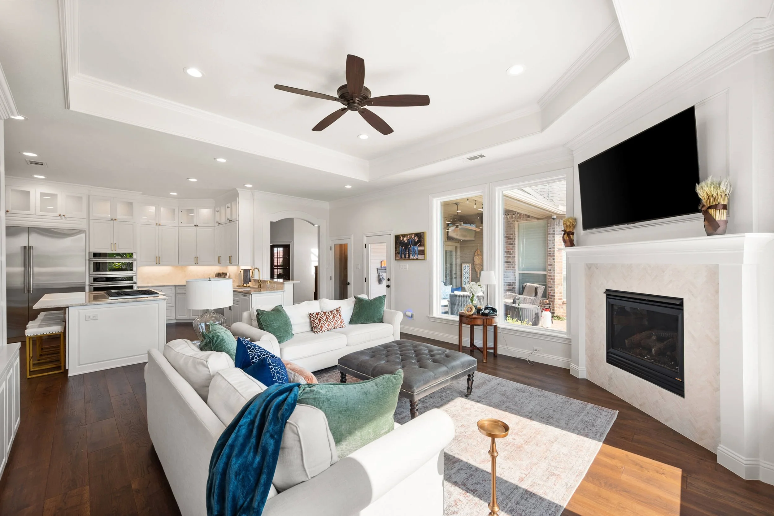 Living room with white sofa, green and patterned throw pillows, dark wood flooring, fireplace, mounted TV, ceiling fan, adjacent to kitchen with white cabinets, stainless steel appliances, and a view of outdoor patio.