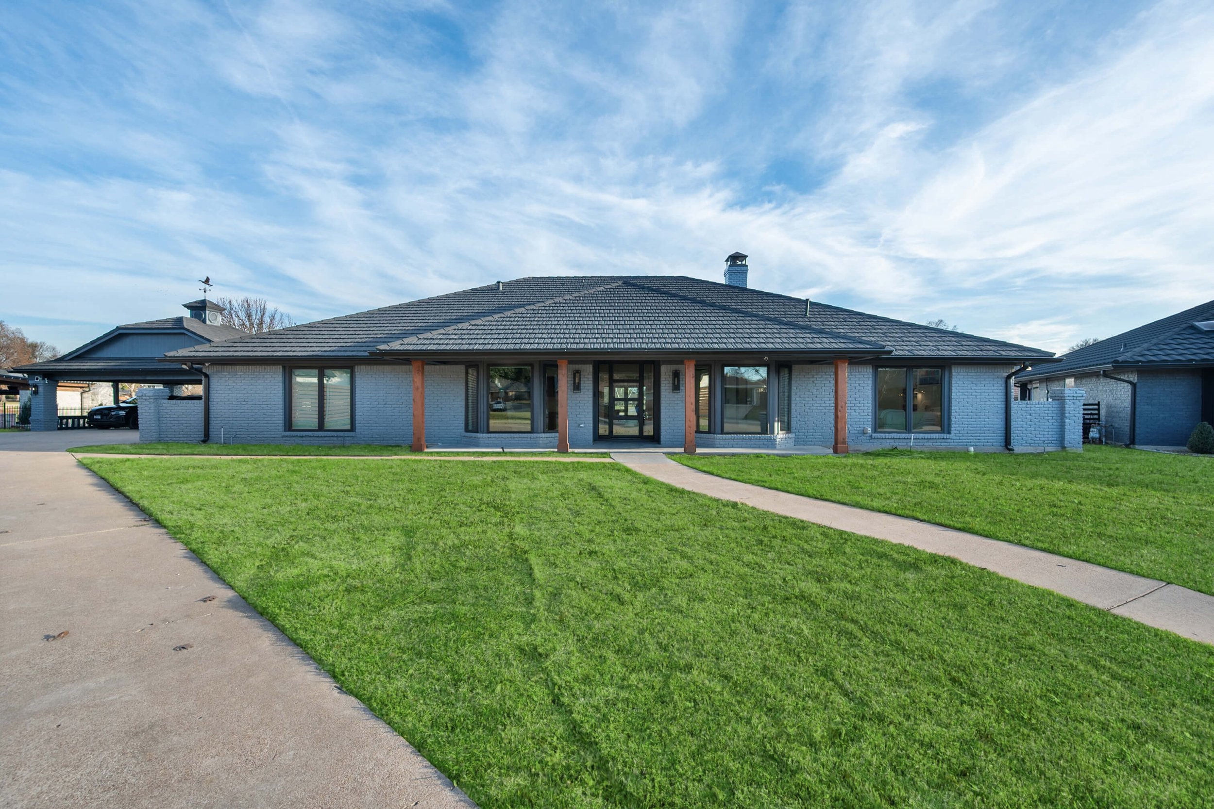 Front view of a single-story house with a dark tiled roof, light-colored brick walls, wooden support beams, large windows, a freshly mowed lawn, and a concrete walkway leading to the entrance, under a partly cloudy sky built by Lakeland Custom Homes.