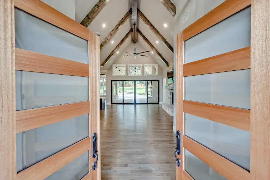 Open wooden and glass sliding doors reveal a spacious living room with vaulted ceiling, exposed wooden beams, a ceiling fan, a large fireplace, and sliding glass doors leading outside.