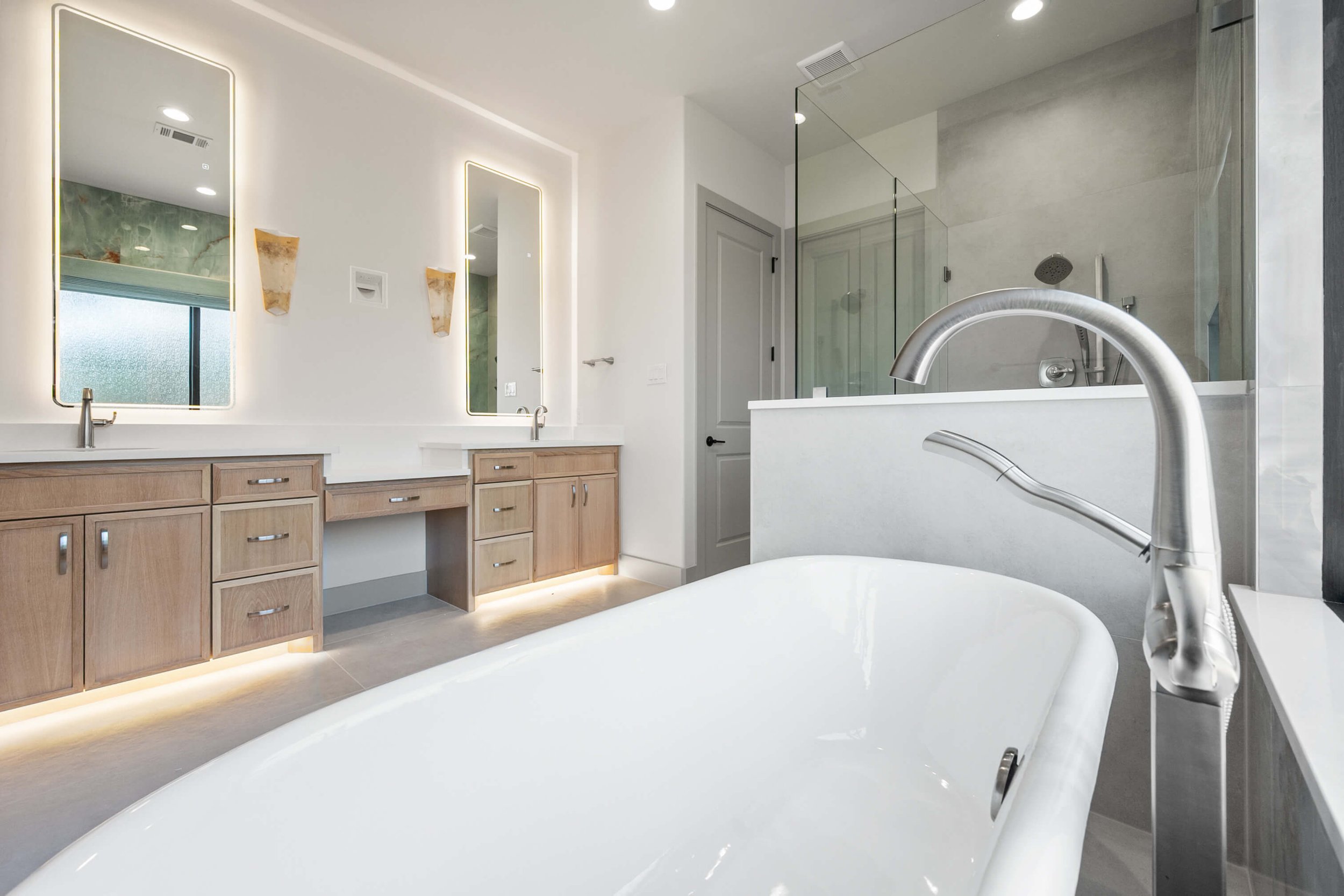 Modern bathroom with double vanity sinks, large mirrors with backlit LED lighting, wooden cabinets, a free-standing bathtub with a chrome faucet, and a walk-in glass shower with a rain showerhead built by Lakeland Custom Homes.