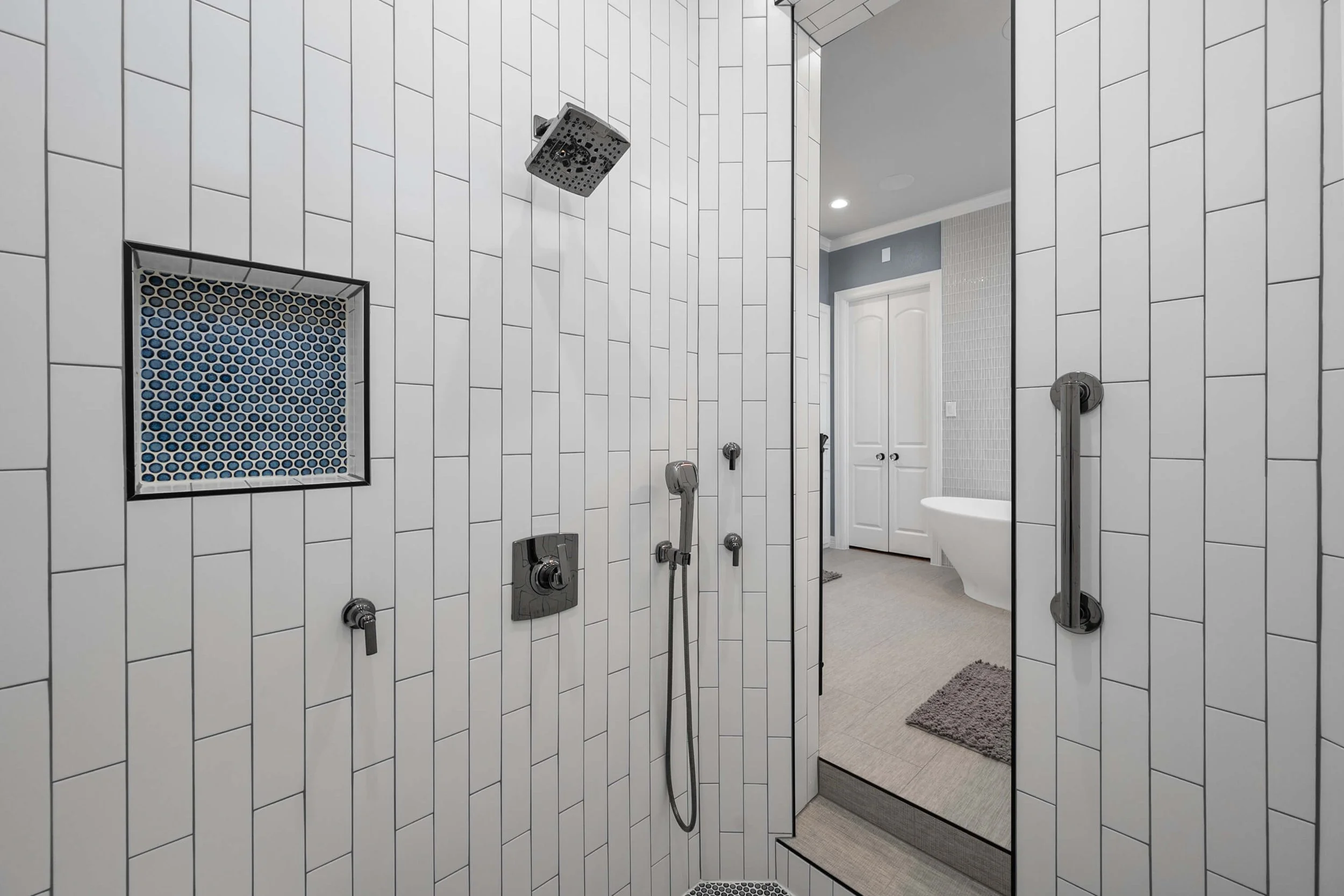 Modern walk-in shower with white rectangular tiles, a rainfall showerhead, handheld shower, and a recessed soap niche; steps down to a beige bathroom with a white bathtub and double doors in the background.