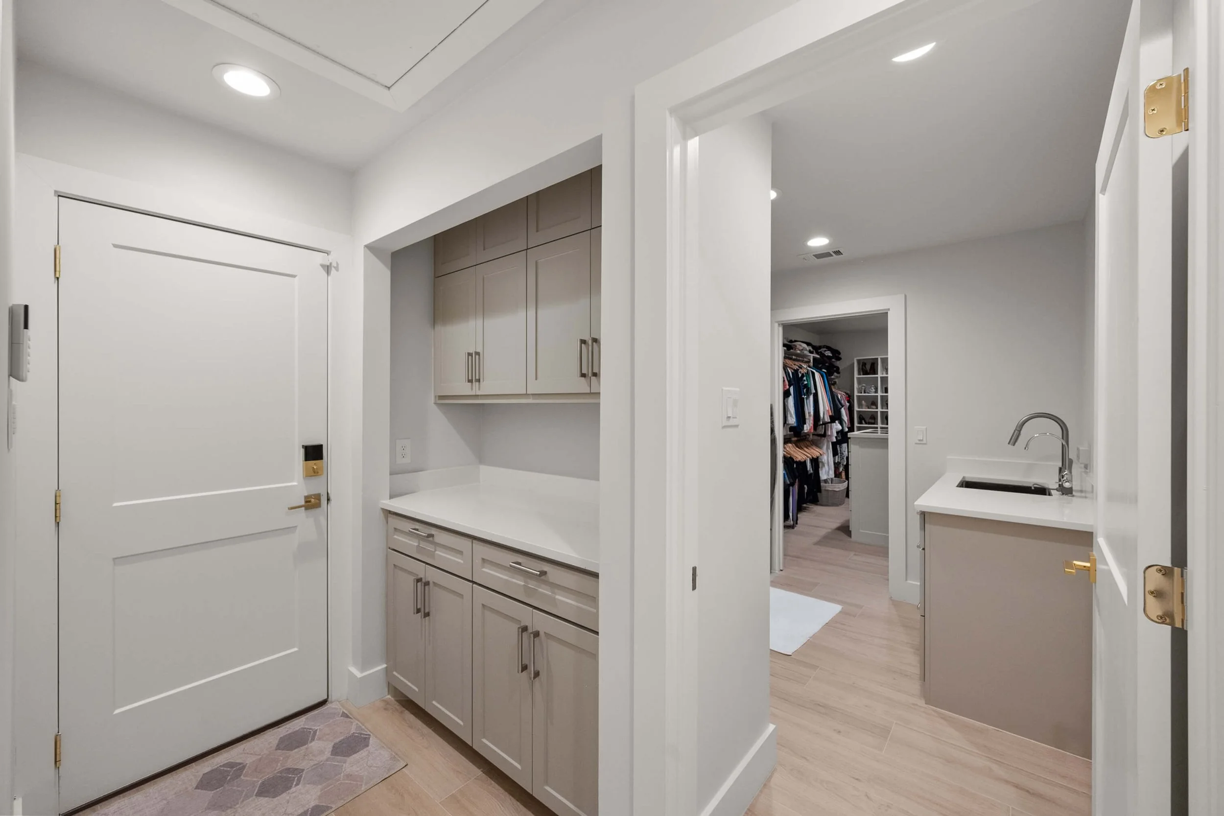 Interior view of a laundry or utility room with beige cabinetry, a white countertop, a small sink, and an open doorway leading to a walk-in closet with clothes and shelves built by Lakeland Custom Homes.
