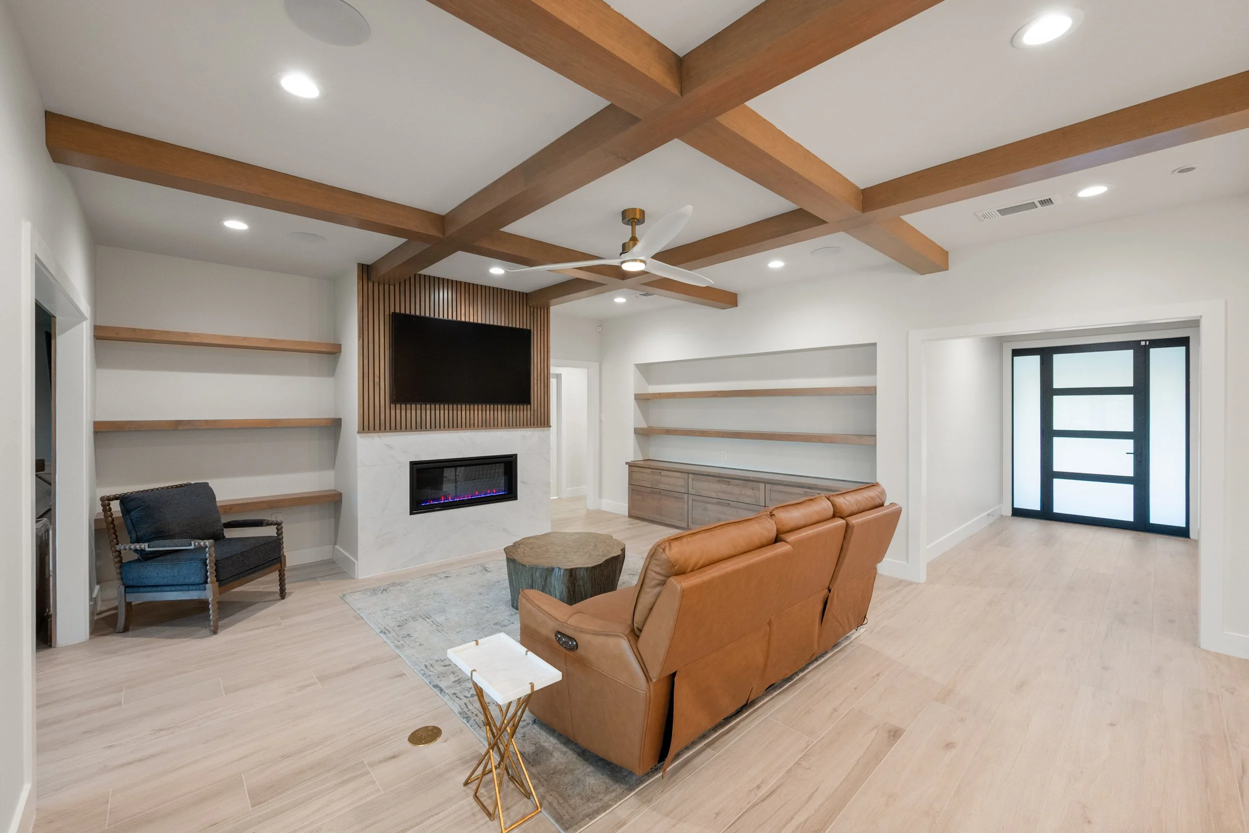 Modern living room with a tan leather sofa, a black armchair, a wood slab coffee table, built-in shelves, a wall-mounted TV, a fireplace, wood ceiling beams, recessed lighting, and a black front door with frosted glass panels.