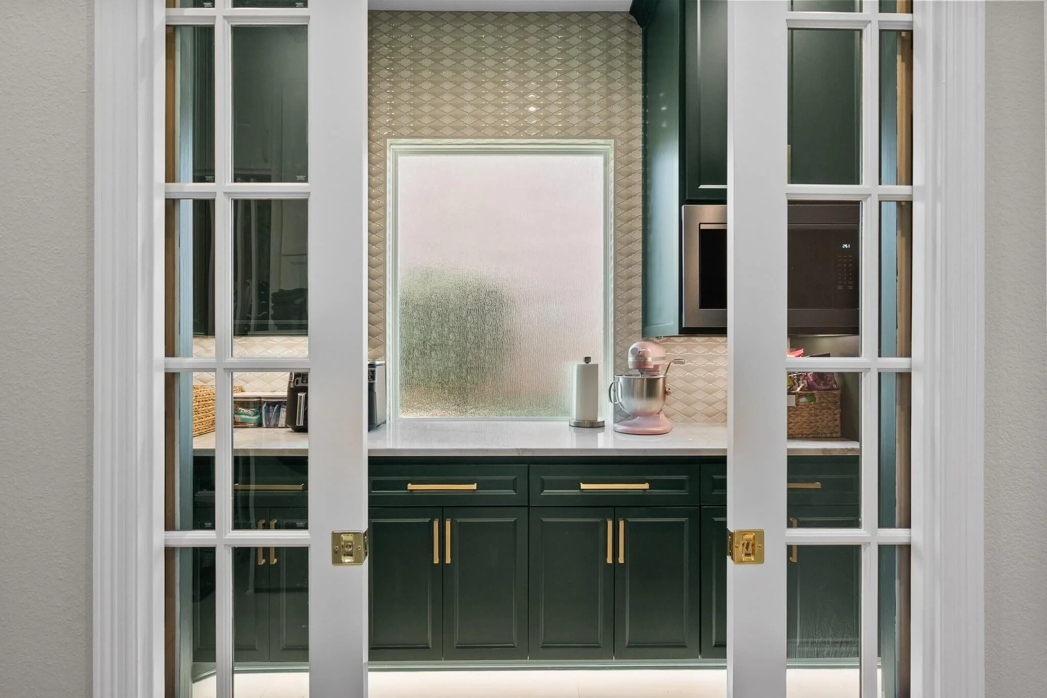 Kitchen viewed through white glass-paneled sliding doors, featuring green cabinets, a textured cream backsplash, a frosted window, a stand mixer, and small appliances on the countertop.