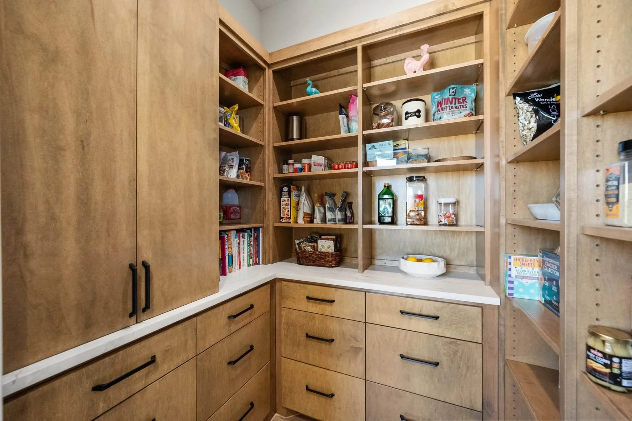 Empty wooden pantry with open shelves filled with snacks, condiments, books, and kitchen items. Cabinets and drawers with black handles.