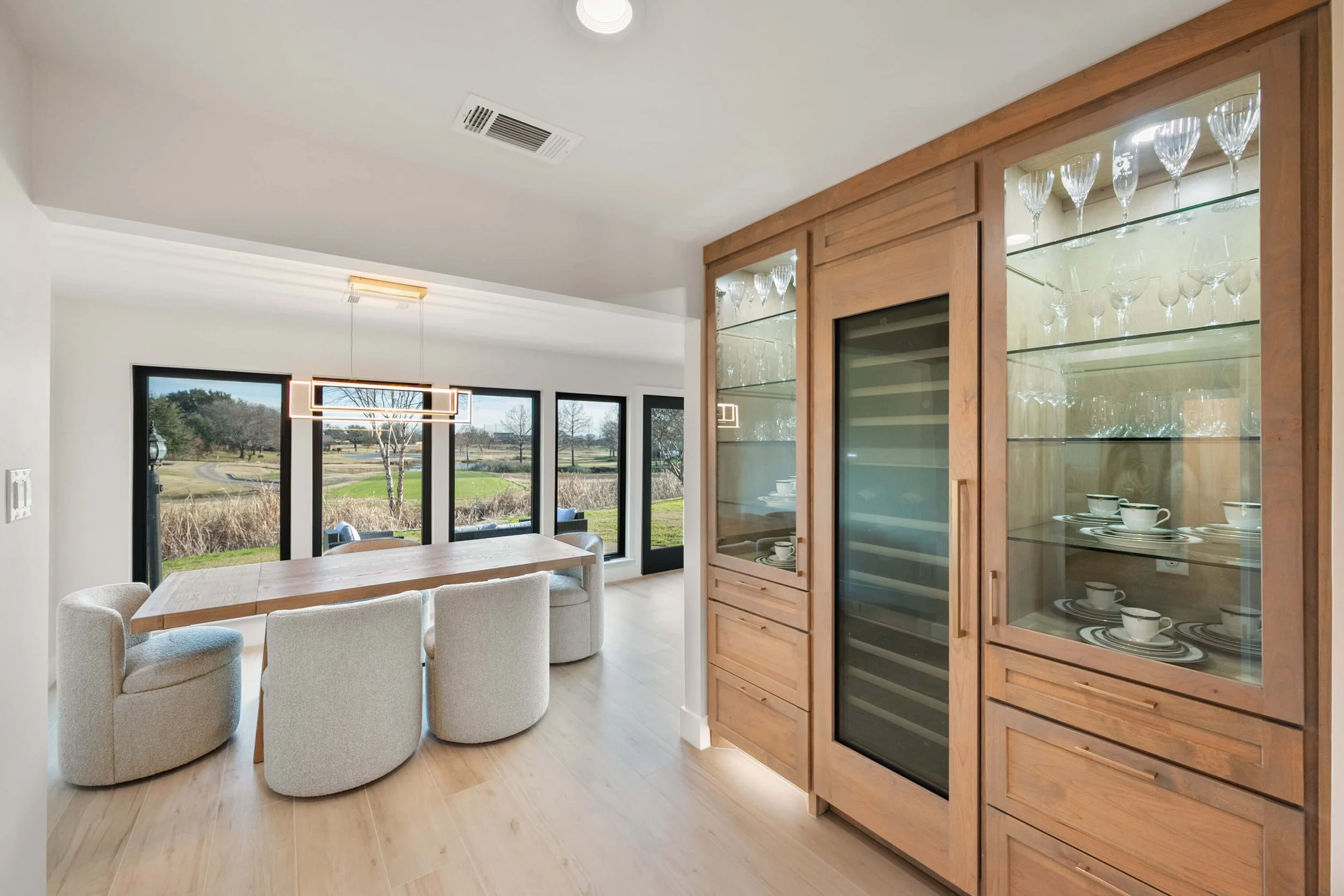 Dining area with a large wooden table, four upholstered chairs, and a view of outdoors through large windows, with built-in glassware and dish storage cabinets.