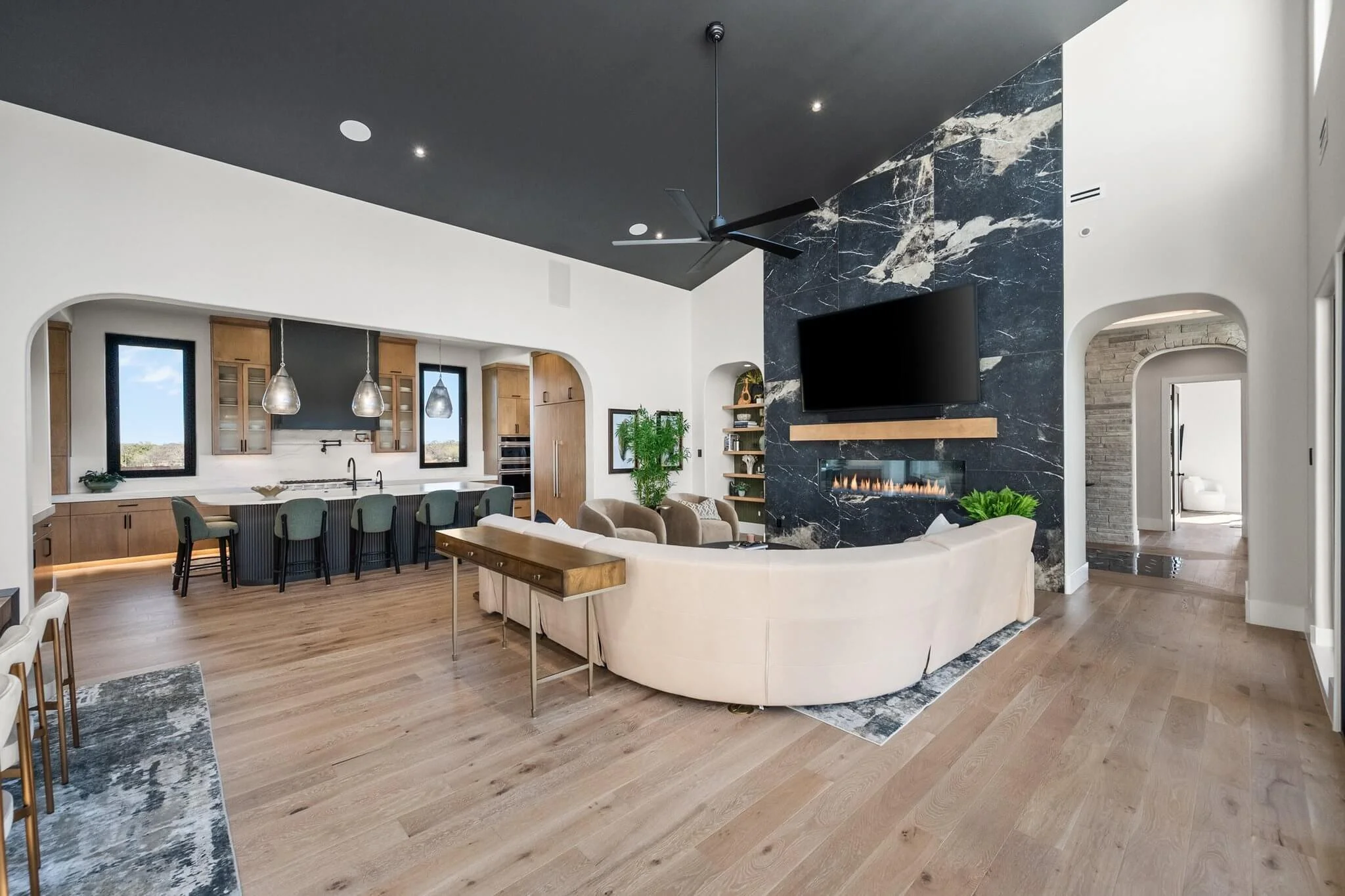 Modern open-concept living room with a black ceiling, light wood flooring, a large beige curved sofa, a black marble accent wall with a mounted TV and a linear fireplace, a ceiling fan, and a kitchen area with pendant lights and bar stools.