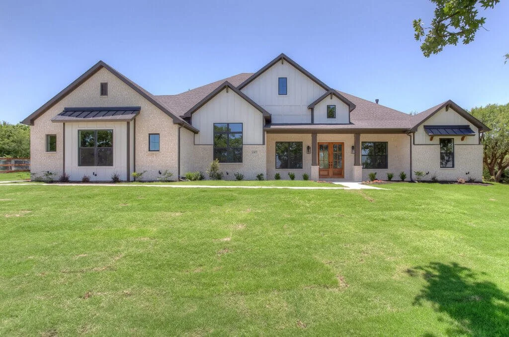 Modern house with white brick and vertical siding, large windows, and a wooden front door, surrounded by a green lawn and some trees in Mansfield, TX.