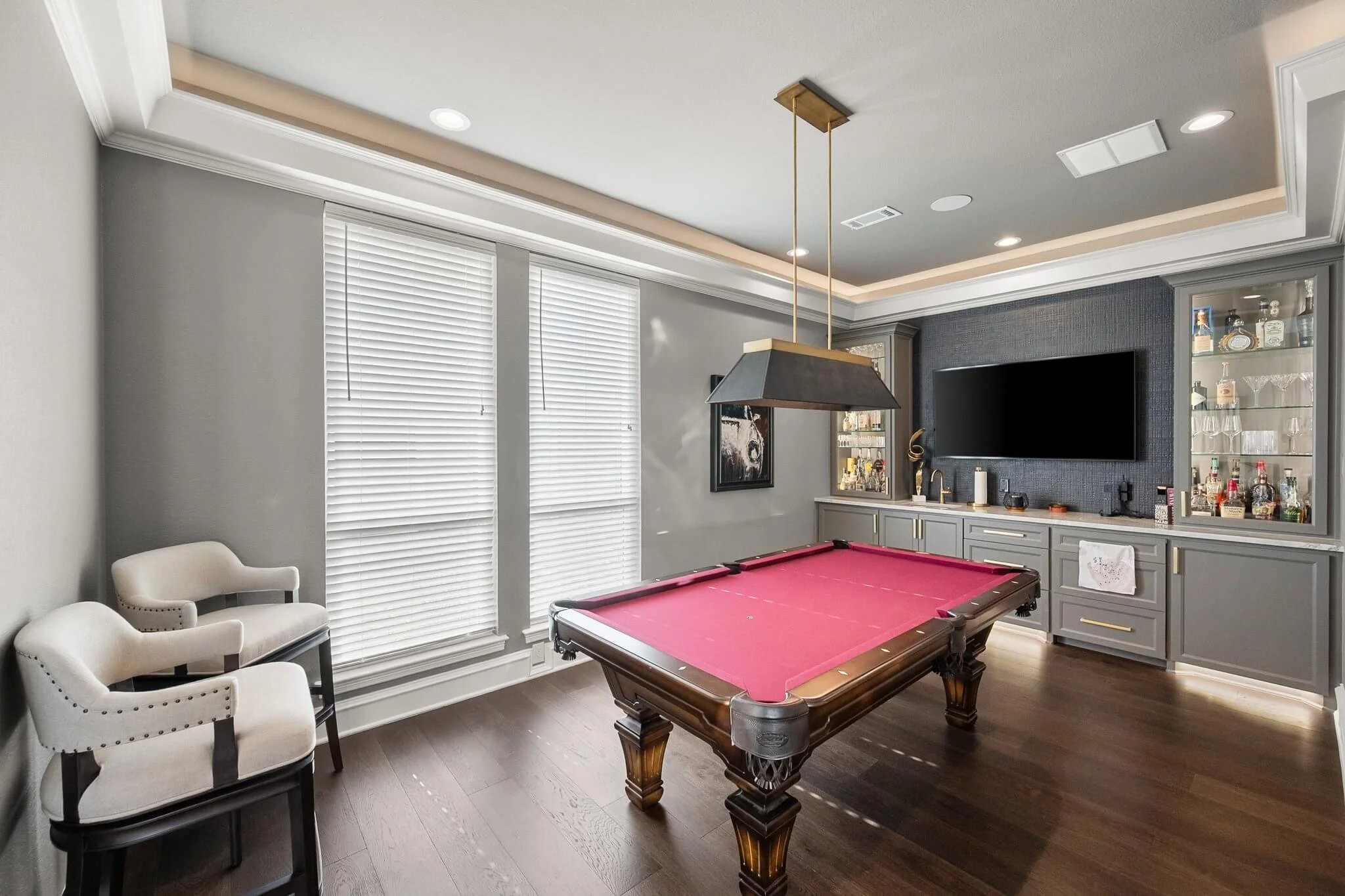 A game room featuring a pool table with a red felt surface, two white armchairs, large windows with blinds, a built-in bar with glass shelves, a flat-screen TV, and a ceiling light fixture in Mansfield, TX.