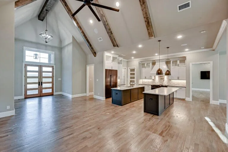 Open-concept kitchen and living area in a modern home with hardwood floors, a vaulted ceiling with exposed wooden beams, and large front door with glass panels.