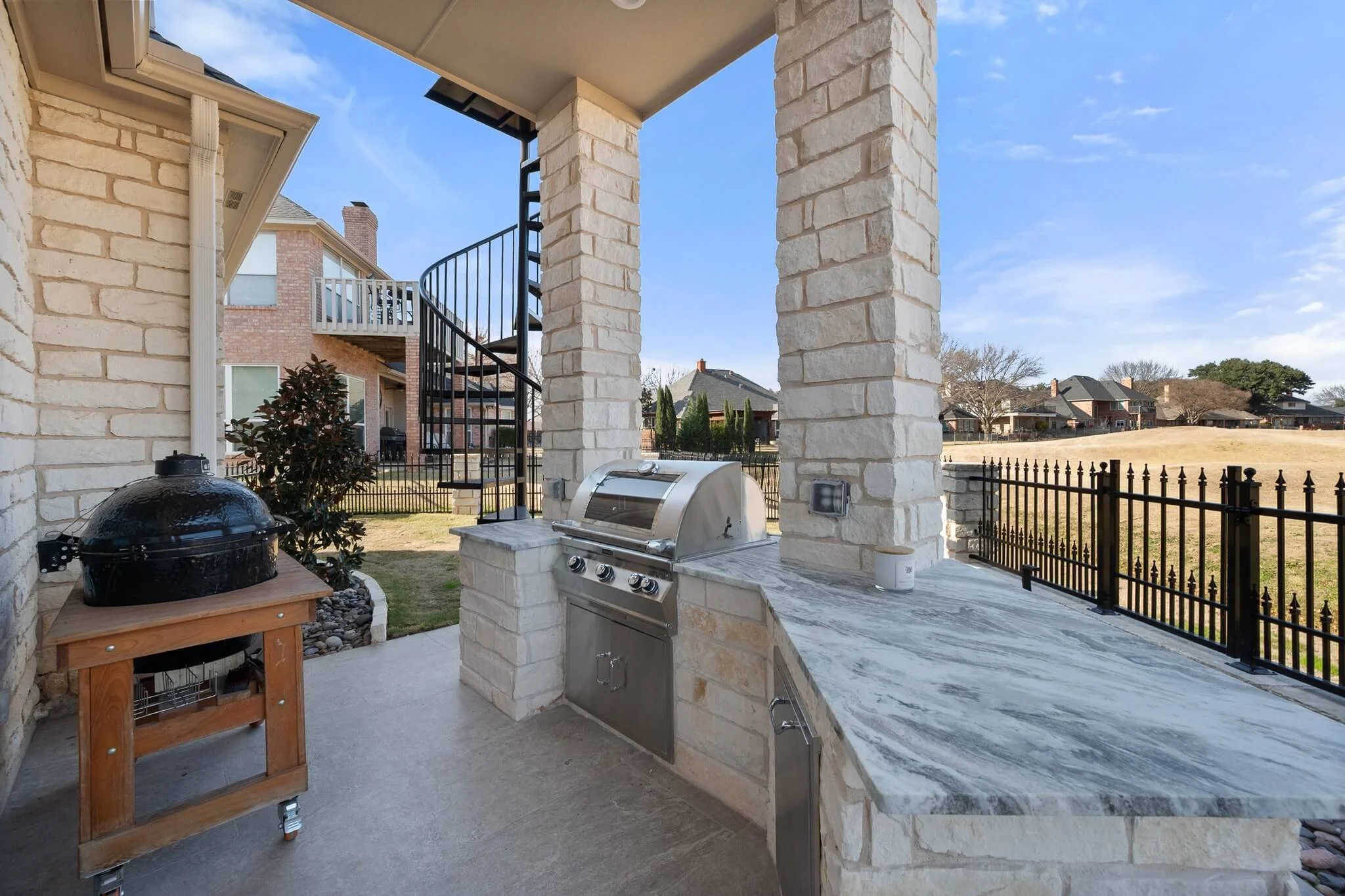 Outdoor patio area with a built-in stainless steel grill, a black kettle-style smoker on a wooden stand, and a marble countertop under a covered porch. In the background, there is a view of neighboring houses, a grassy yard, and a black metal fence.