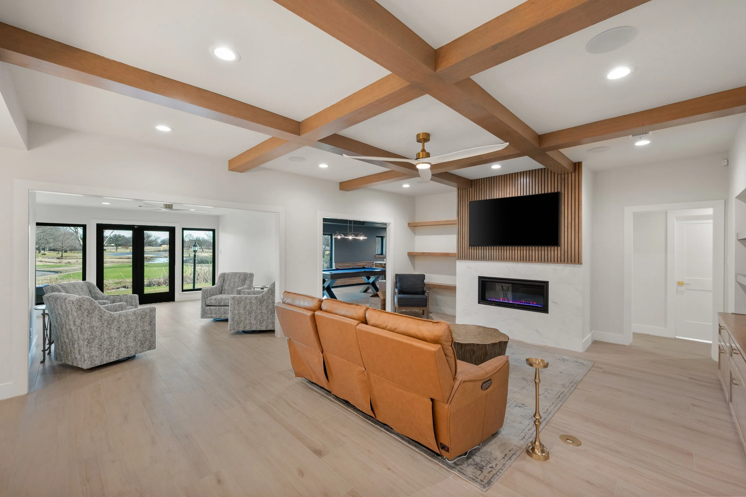 Living room with modern decor, tan leather sofa, gray armchairs, fireplace, mounted TV, wooden ceiling beams, and natural light from large windows built by Lakeland Custom Homes.