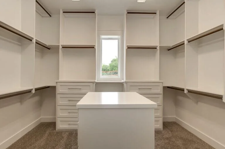 Empty walk-in closet with built-in shelves, drawers, and a central island, illuminated by natural light from a small window.