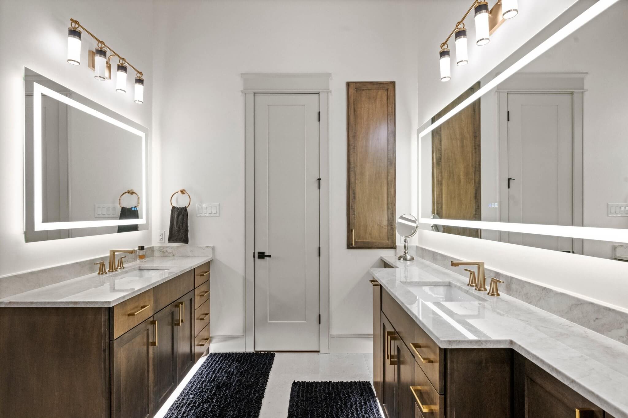 Modern bathroom with dual vanities, white marble countertops, gold fixtures, illuminated mirrors, and a wooden cabinet between the doors  built by Lakeland Custom Homes..