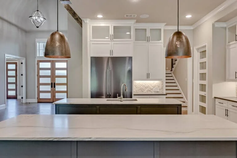 Modern kitchen with white cabinets, stainless steel refrigerator, marble island, pendant lights, and staircase in the background.