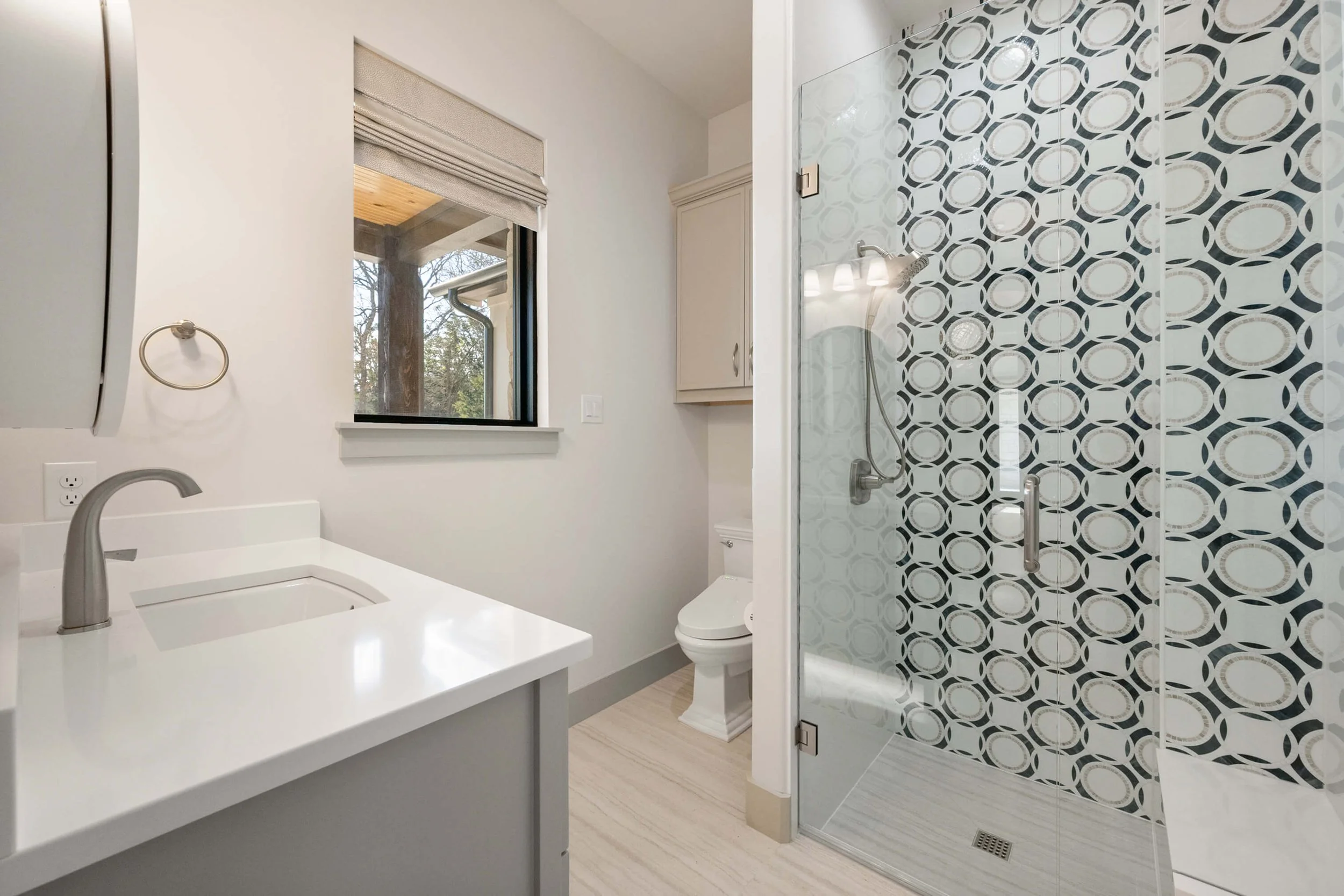 Bathroom with white countertop sink, a window with a view of trees outside, a toilet, and a glass-enclosed shower with patterned tile walls.