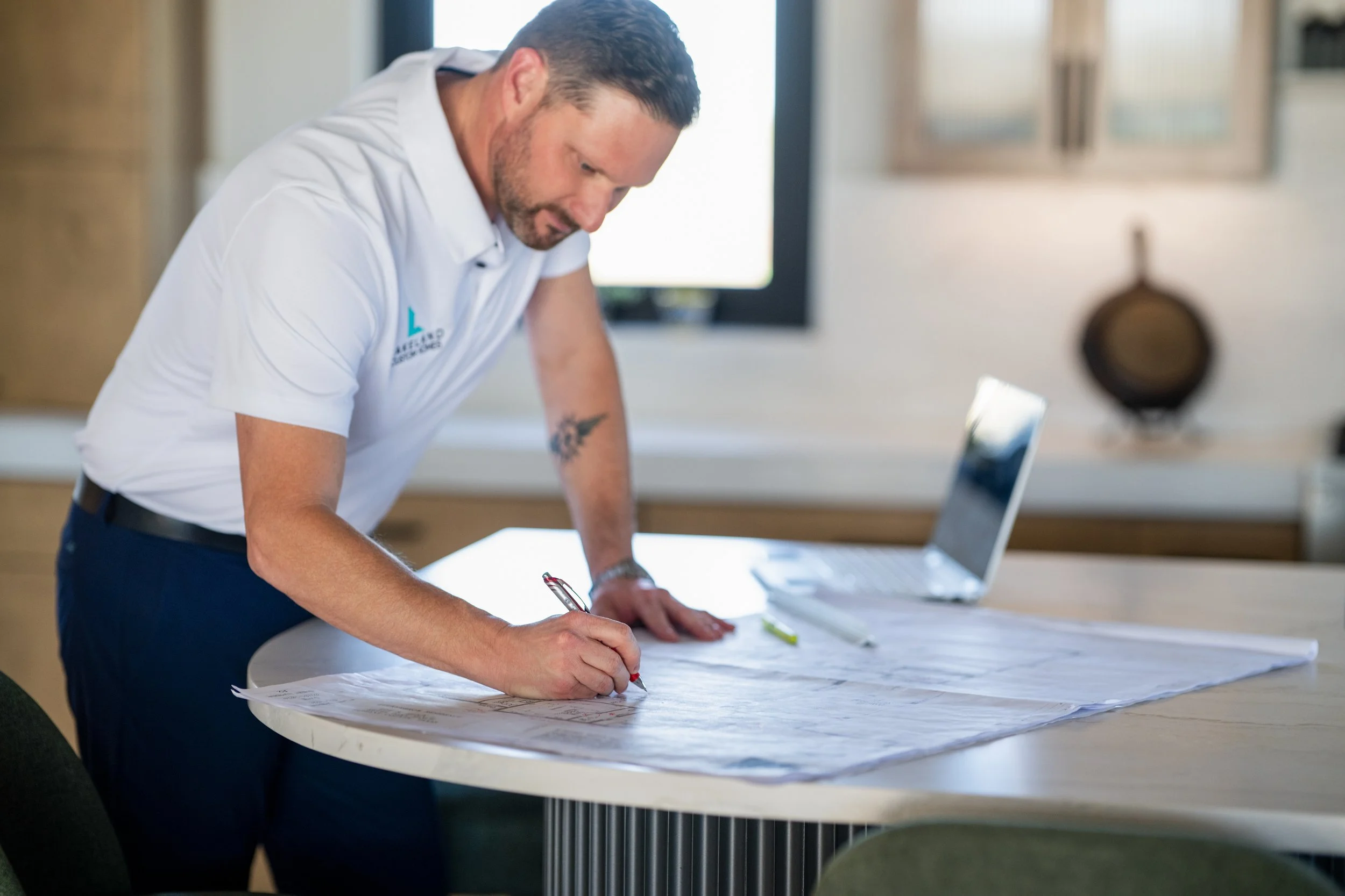A man in a white polo shirt leaning over a table and writing on a large sheet of paper, with a laptop and a green highlighter on the table.