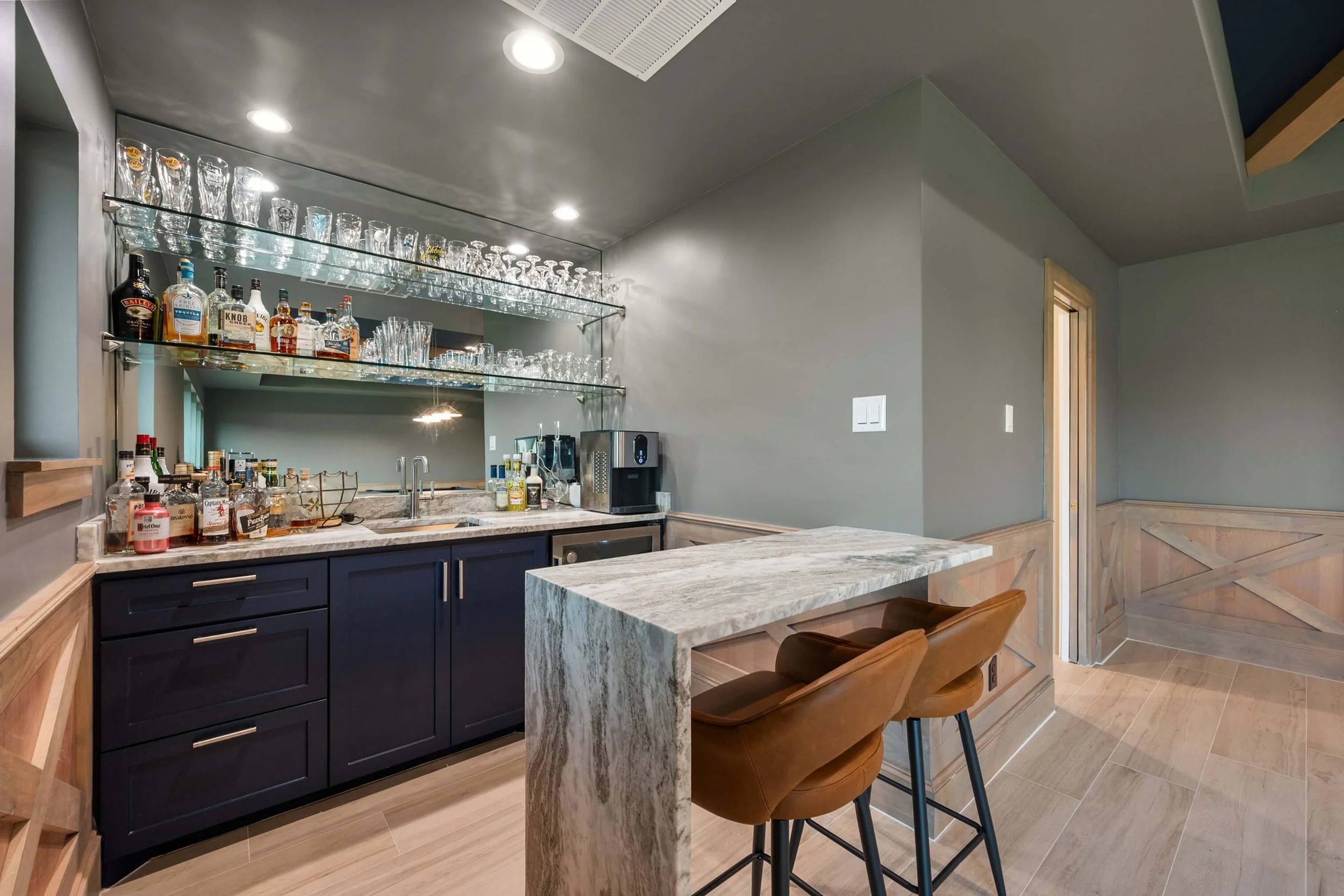 A home bar area with a marble countertop, dark blue lower cabinets, and open glass shelves holding various glassware and bottles of alcohol. Two brown bar stools are in front of the counter, and there is a small sink and a coffee machine in the backg