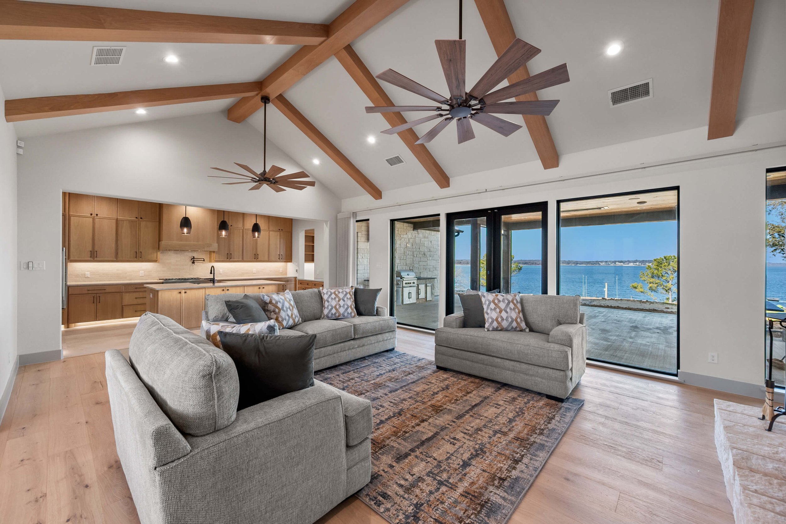 Living room with large sliding glass doors overlooking a lake, wood-beamed ceiling, and modern furnishings including a sofa, armchairs, and a rug.
