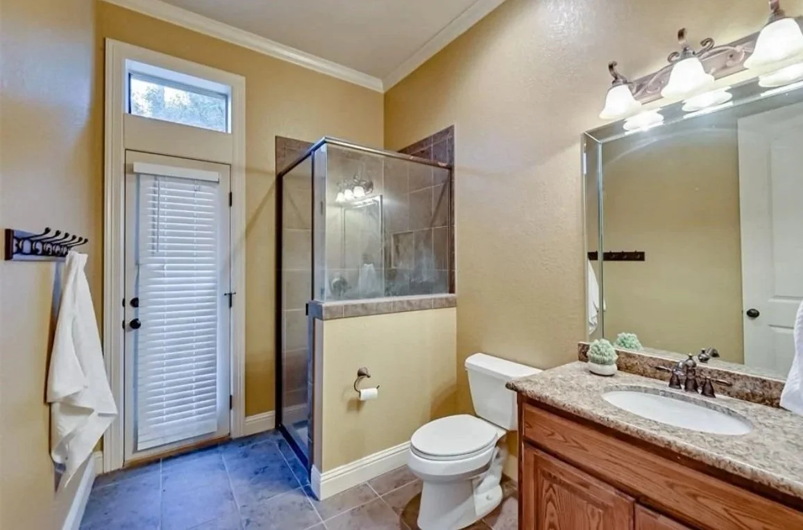 Bathroom with a door and window, a shower stall, a toilet, a wooden vanity with a granite countertop, a large mirror, and wall-mounted light fixtures.