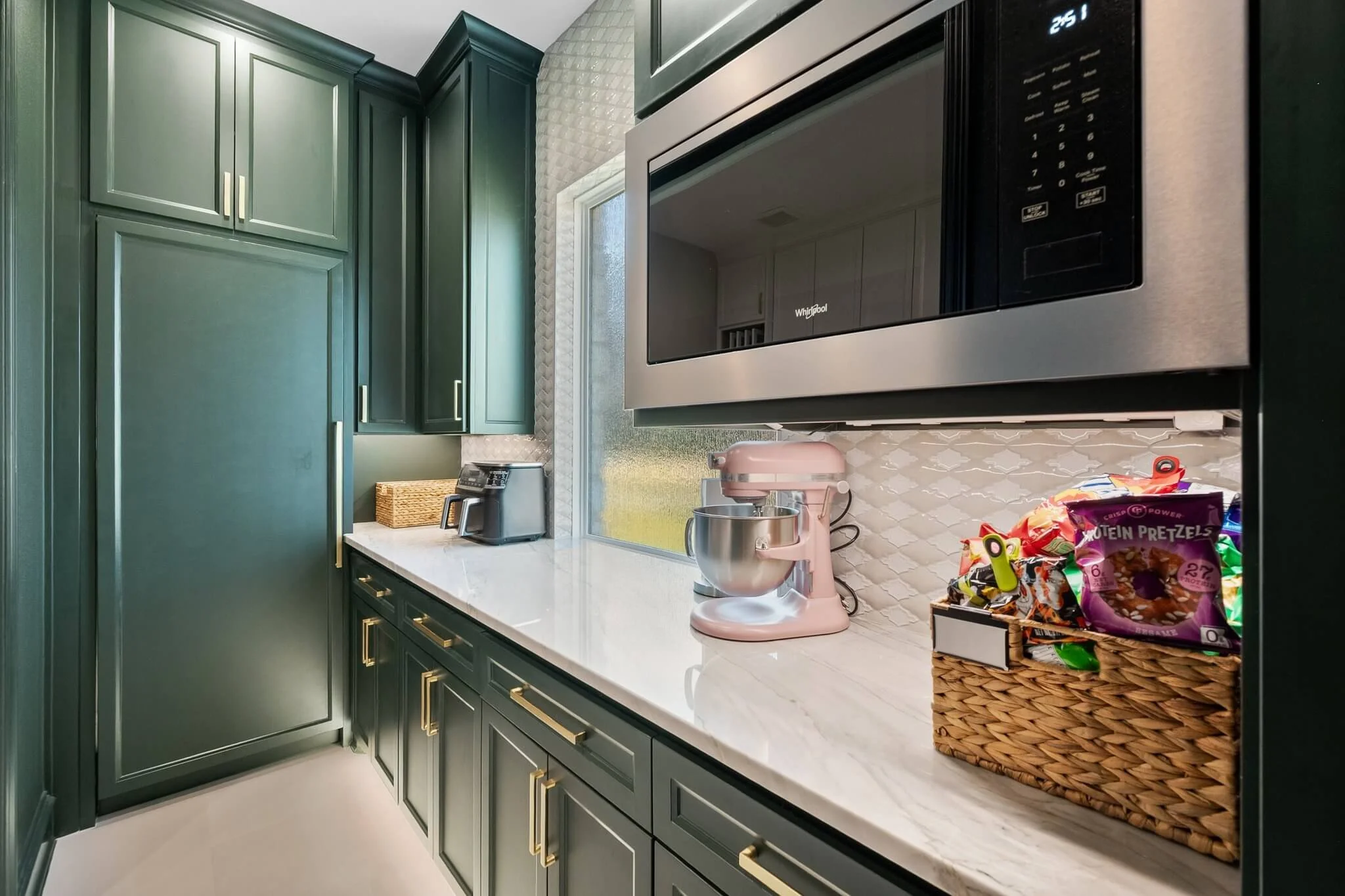 Kitchen countertop with green cabinets, a window, small appliances, a pink stand mixer, and a basket of snacks in Mansfield, TX.