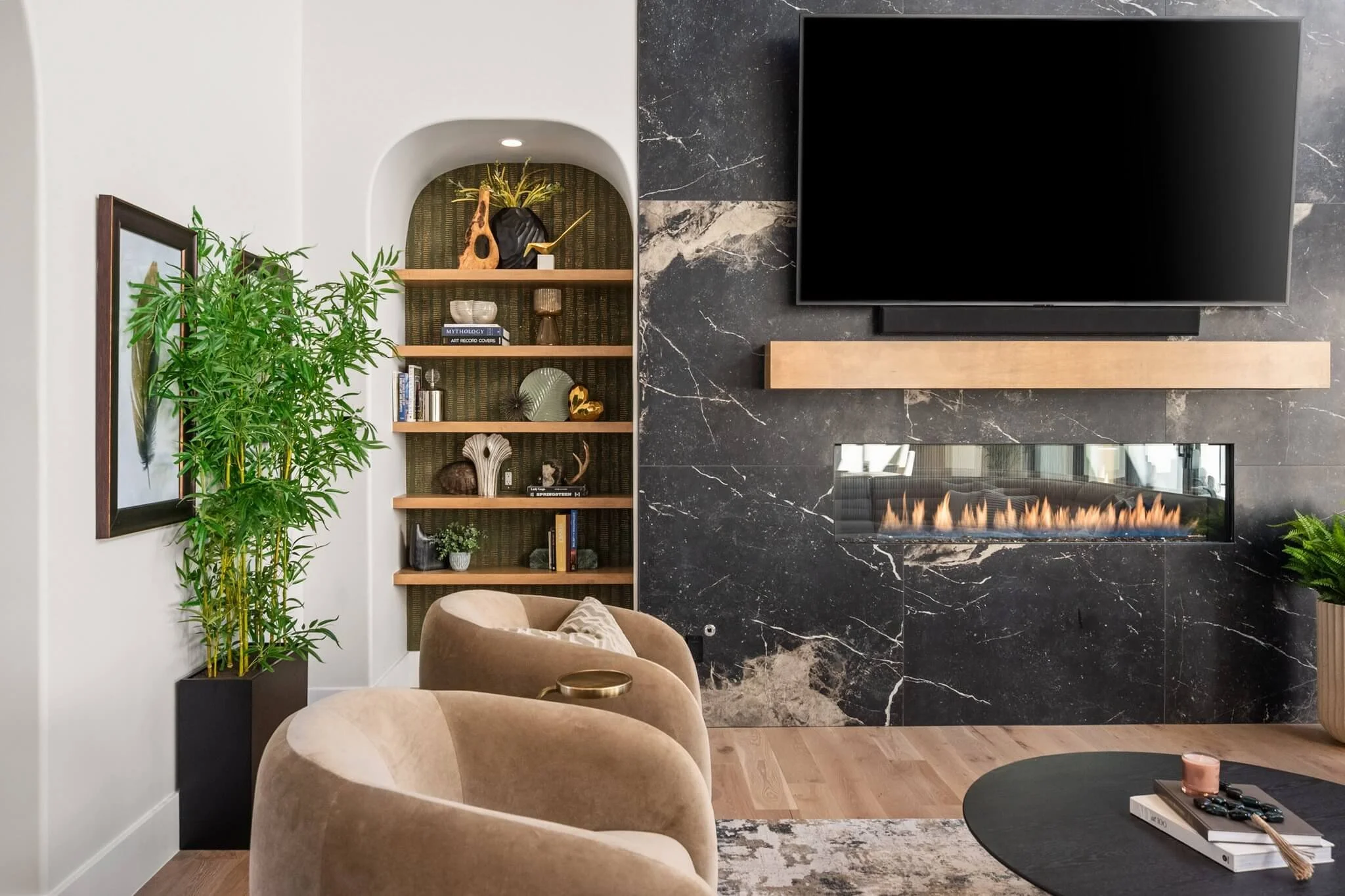 Living room with beige chairs, a black marble fireplace with a TV above, a wooden shelf with decorative items, and a tall green plant in a black planter.