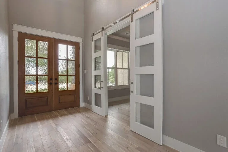 Interior room with wooden floor, double wooden doors with glass panels, and white sliding barn doors leading to an adjacent room with windows