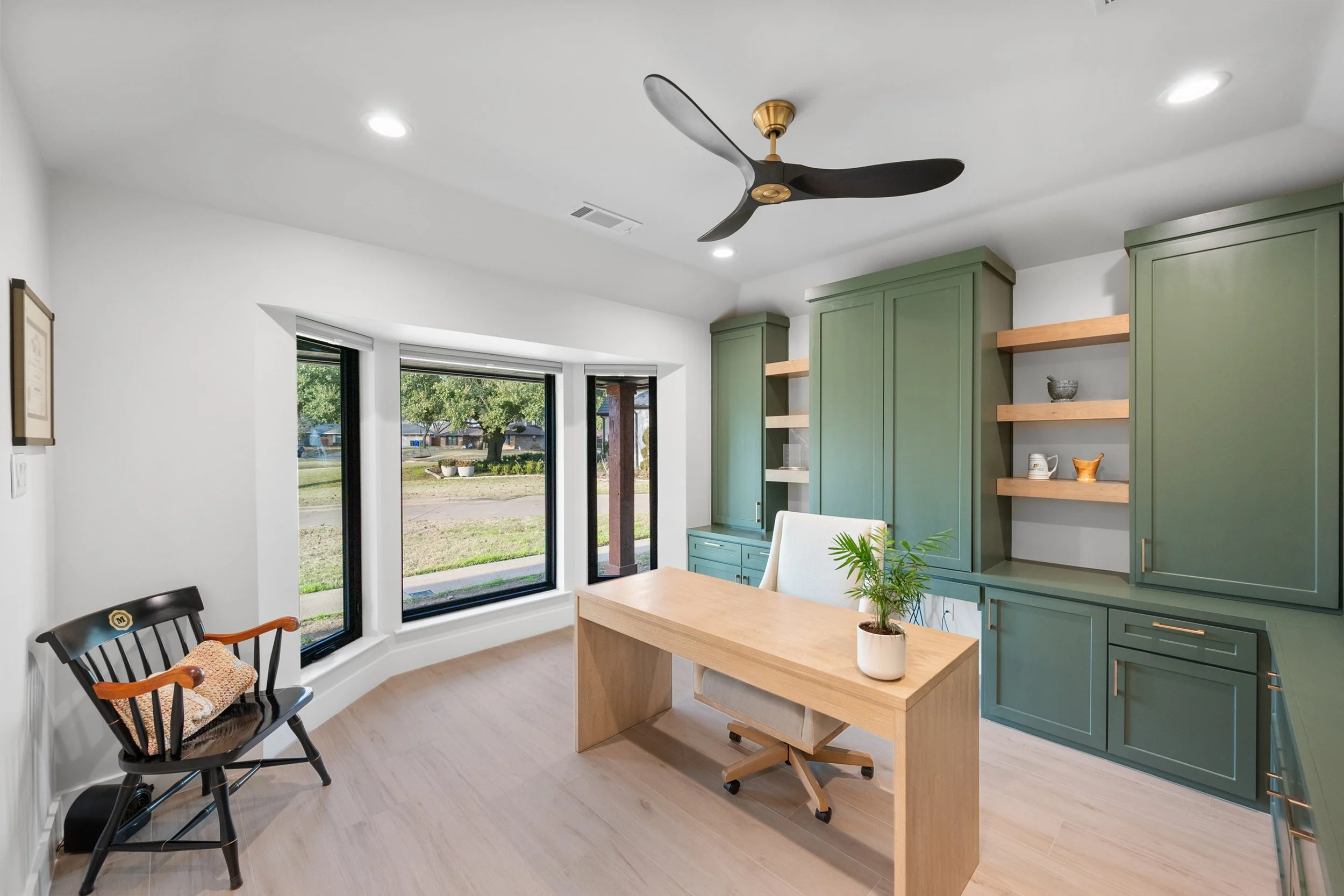 Home office with green built-in cabinets, a wooden desk, a white office chair, a potted plant, and a large window overlooking a yard with trees built by Lakeland Custom Homes.