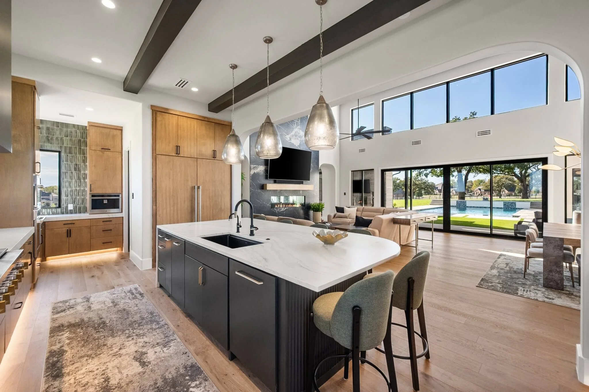 Open-concept kitchen and living room with large windows, wooden cabinets, and modern furnishings, overlooking a backyard with a pool.