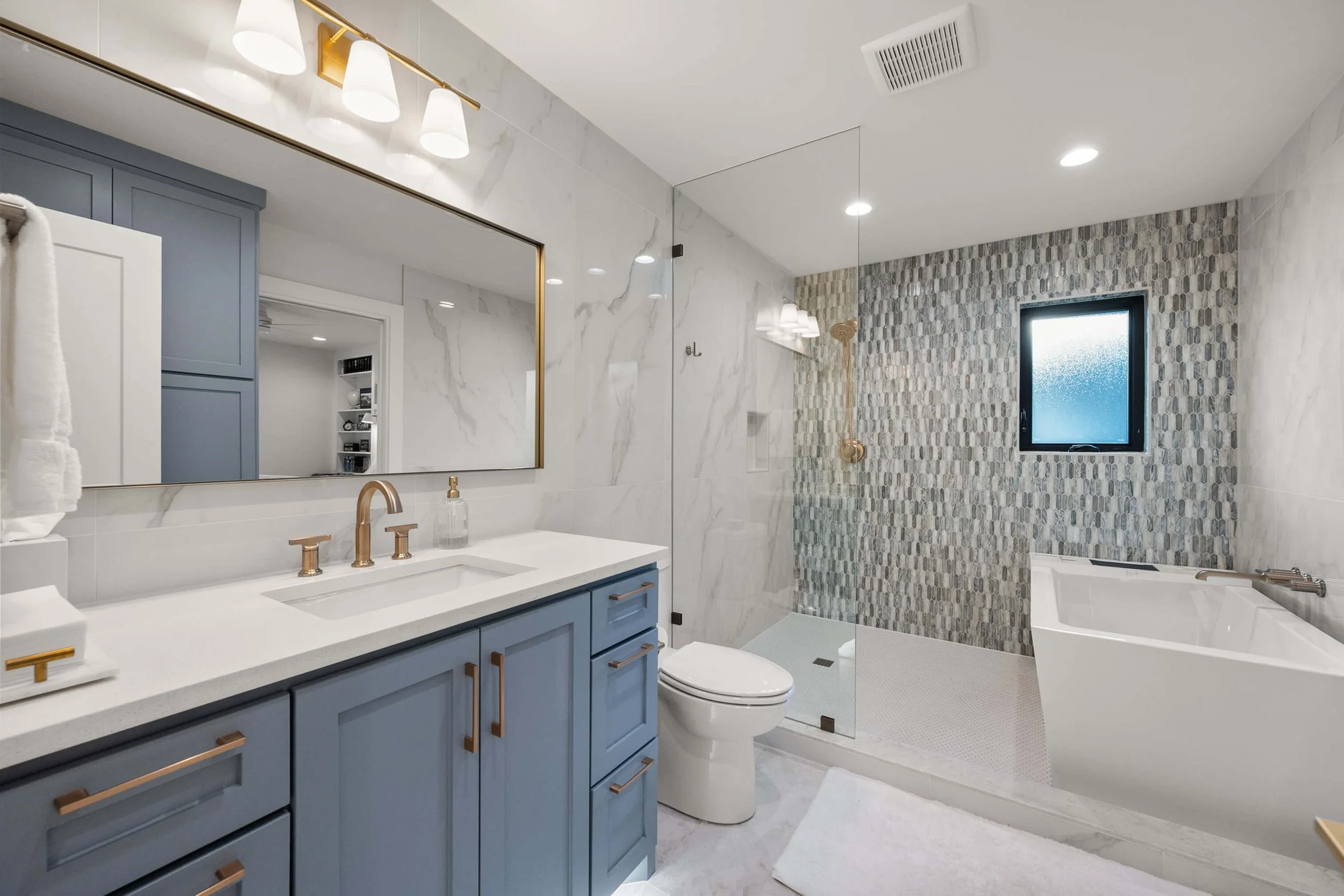 Modern bathroom with blue vanity, large mirror, gold fixtures, walk-in shower with pebble tile, small window, and soaking tub built by Lakeland Custom Homes.