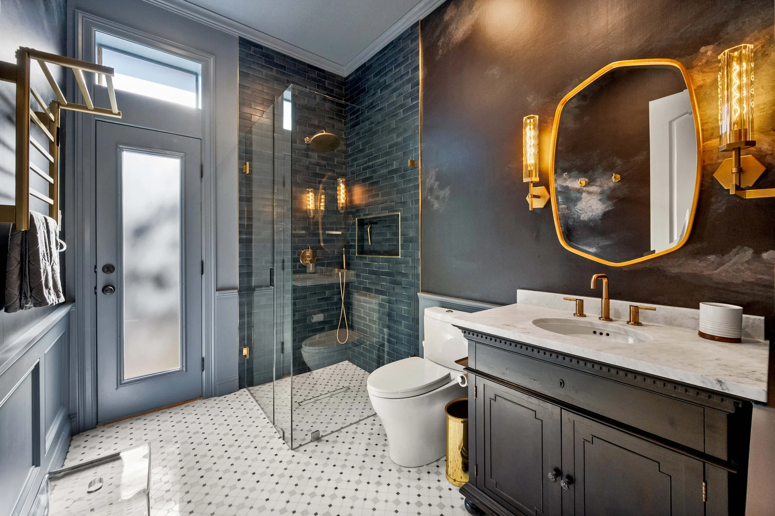 Modern bathroom with dark blue tiled shower, floating vanity with marble countertop, gold fixtures, and a decorative mirror with gold accents. The floor features white hexagonal tiles, and there is a frosted glass door and window.
