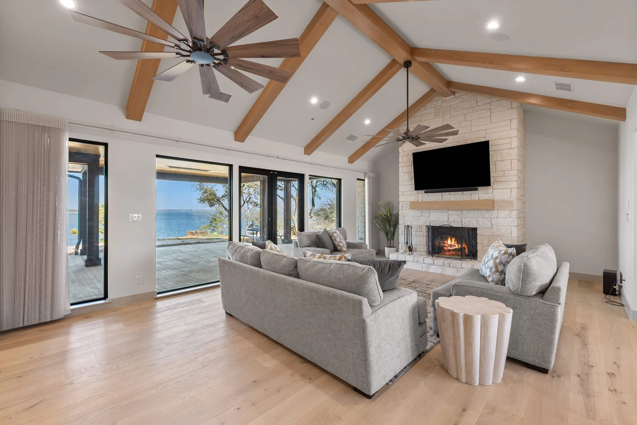 Living room with large windows overlooking water, featuring a stone fireplace with a mounted TV, gray couches, wooden ceiling beams, ceiling fans, and light wood flooring built by Lakeland Custom Homes.