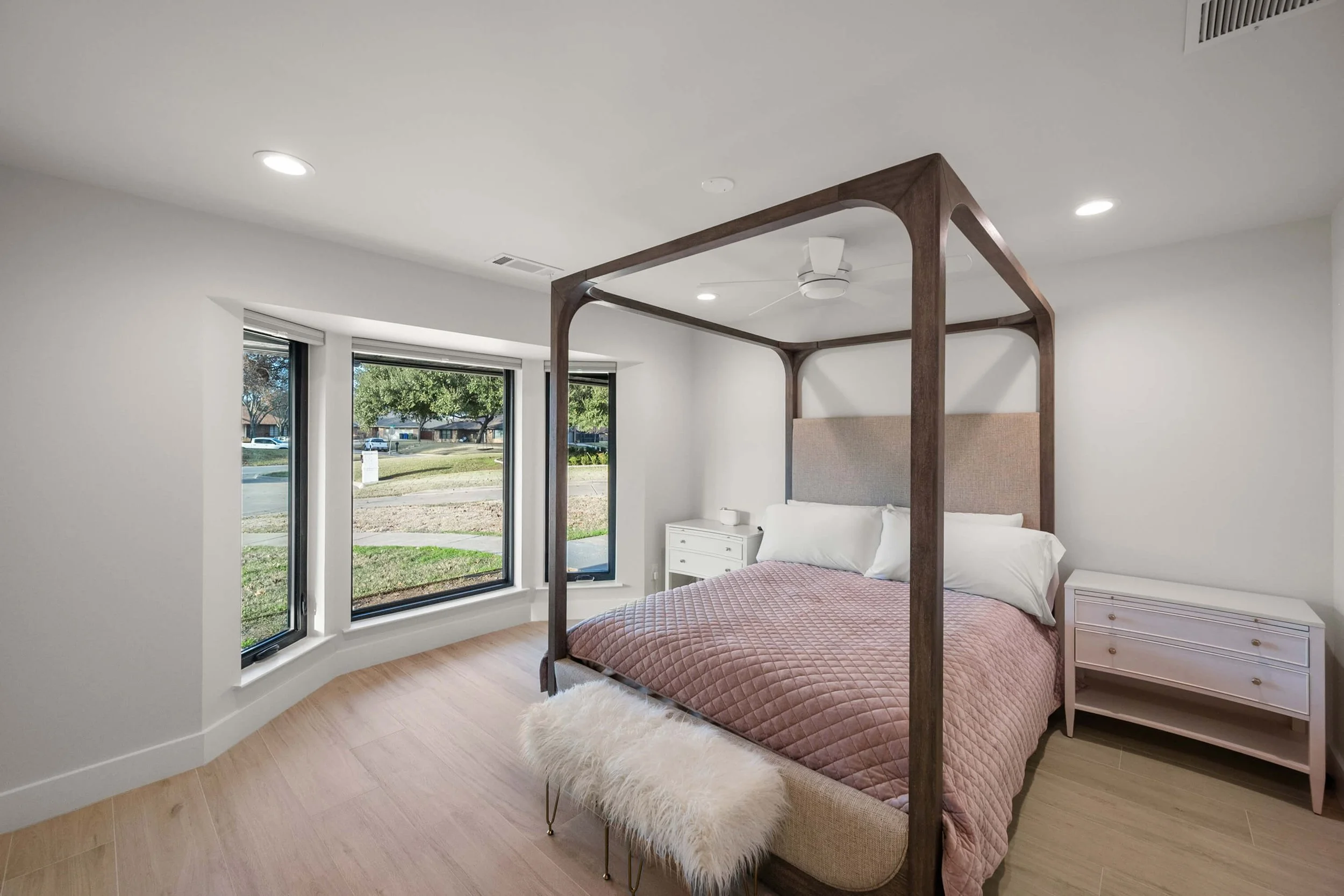 Modern bedroom with a four-poster bed, pink quilt, white pillows, beige headboard, white nightstands, a furry white bench, hardwood floors, and large bay windows with a view of a grassy yard and neighborhood built by Lakeland Custom Homes.