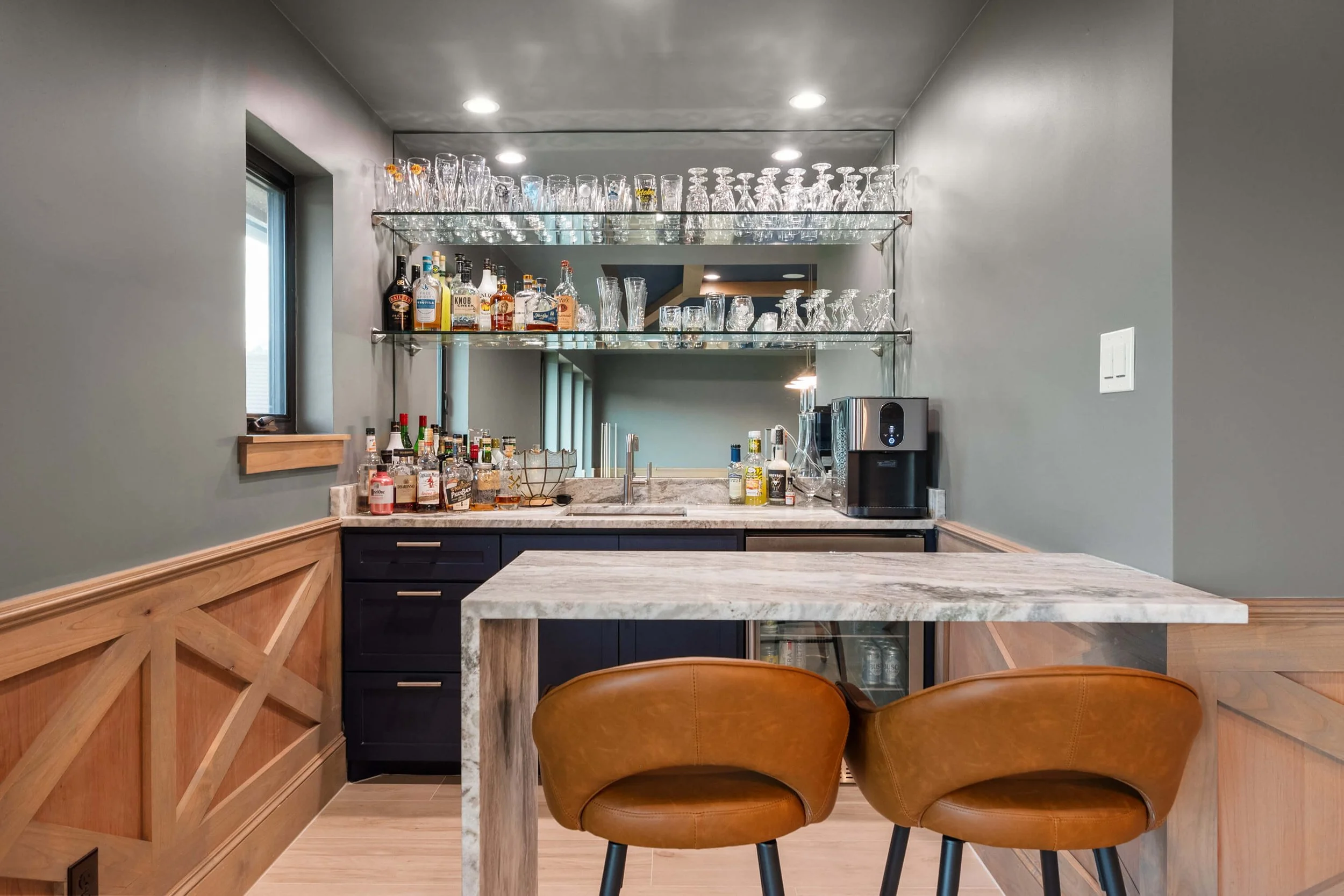 A home bar setup with a gray wall, black cabinets, a marble countertop, open glass shelves with liquor bottles, glassware, and a coffee machine, with two brown leather chairs and a marble-top table in front.