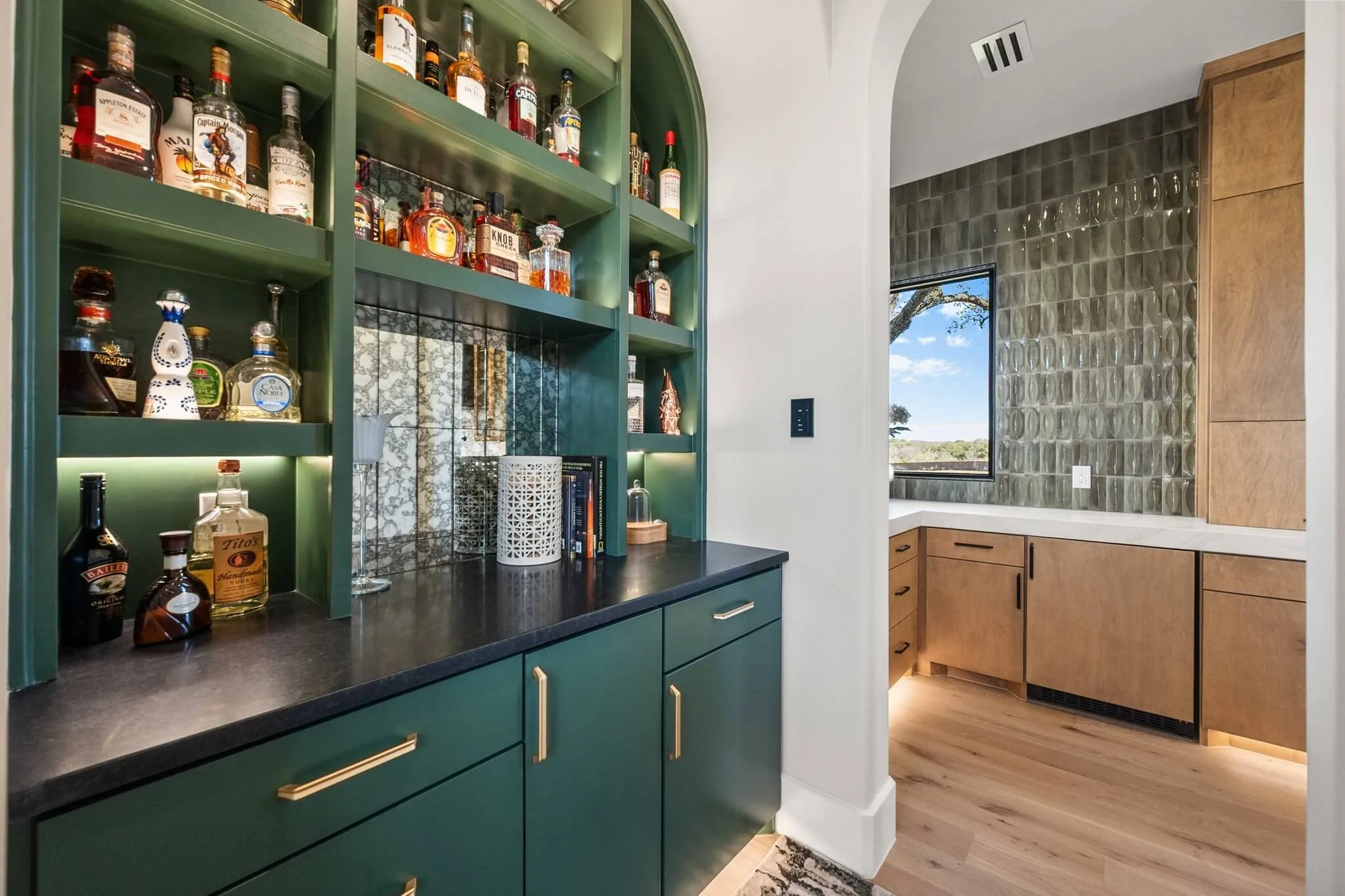 Interior view of a home bar area with green cabinetry and gold handles, displaying various liquor bottles on open shelves. The background shows a kitchen nook with wooden cabinetry, a window, and a textured tile wall.