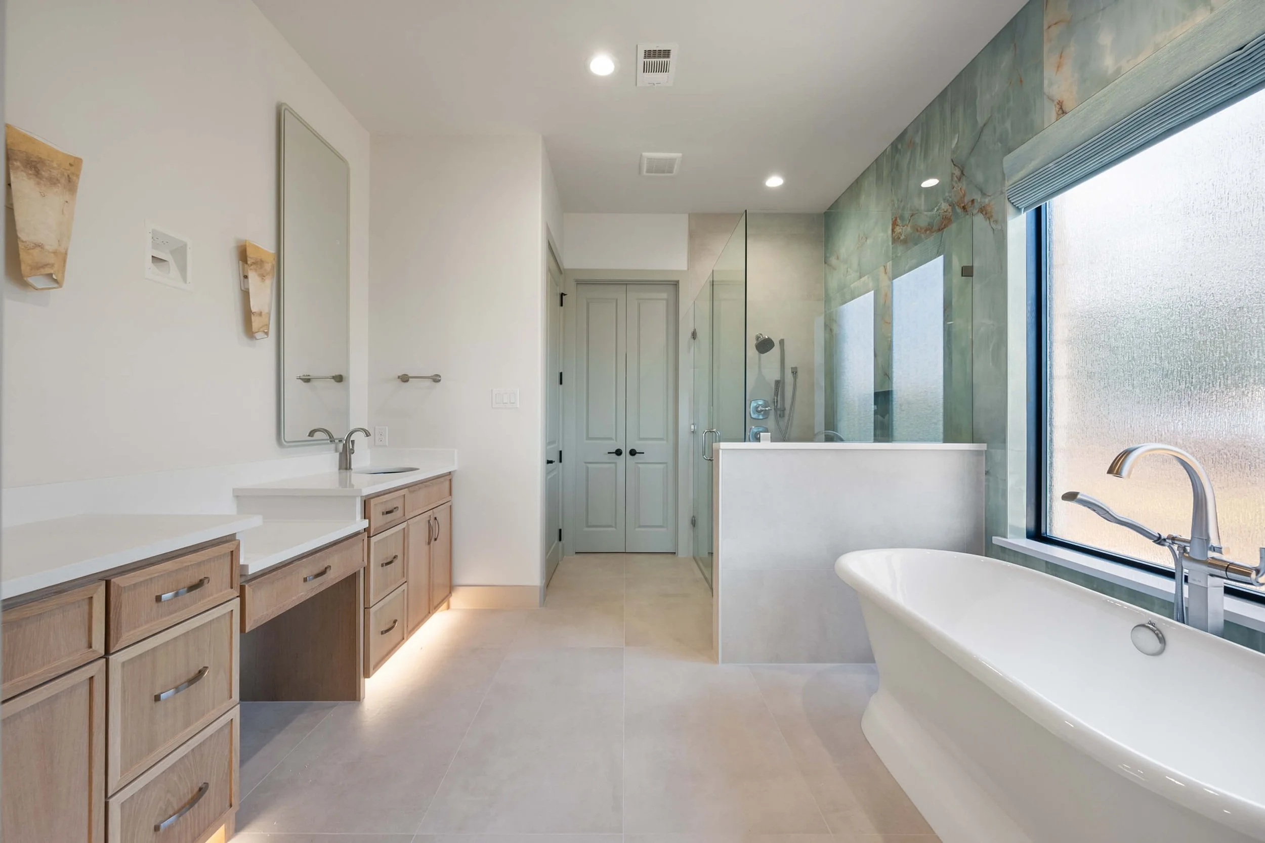 Modern bathroom with wooden cabinets, a large mirror, a freestanding bathtub near a frosted window, and a walk-in shower with glass enclosure and green marble walls.