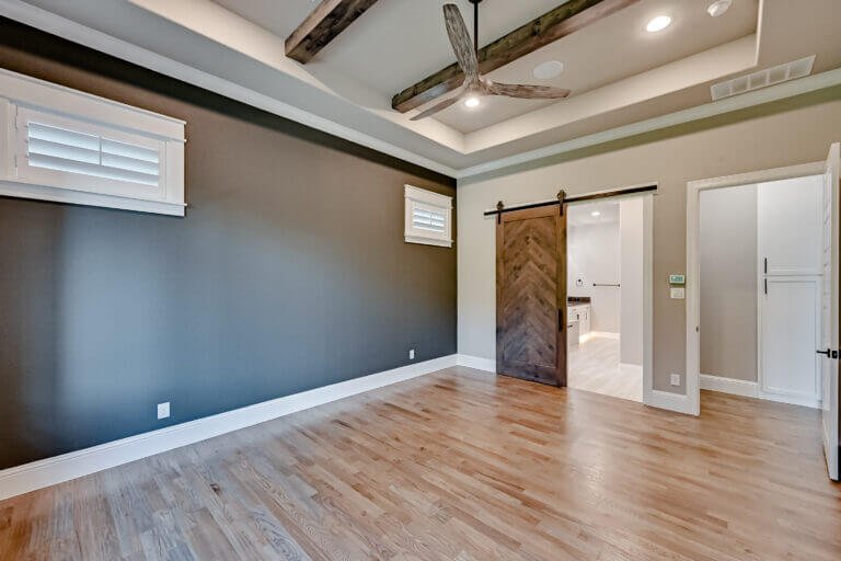 Empty living room with hardwood floors, gray and white walls, and a ceiling with exposed wooden beams and a ceiling fan.